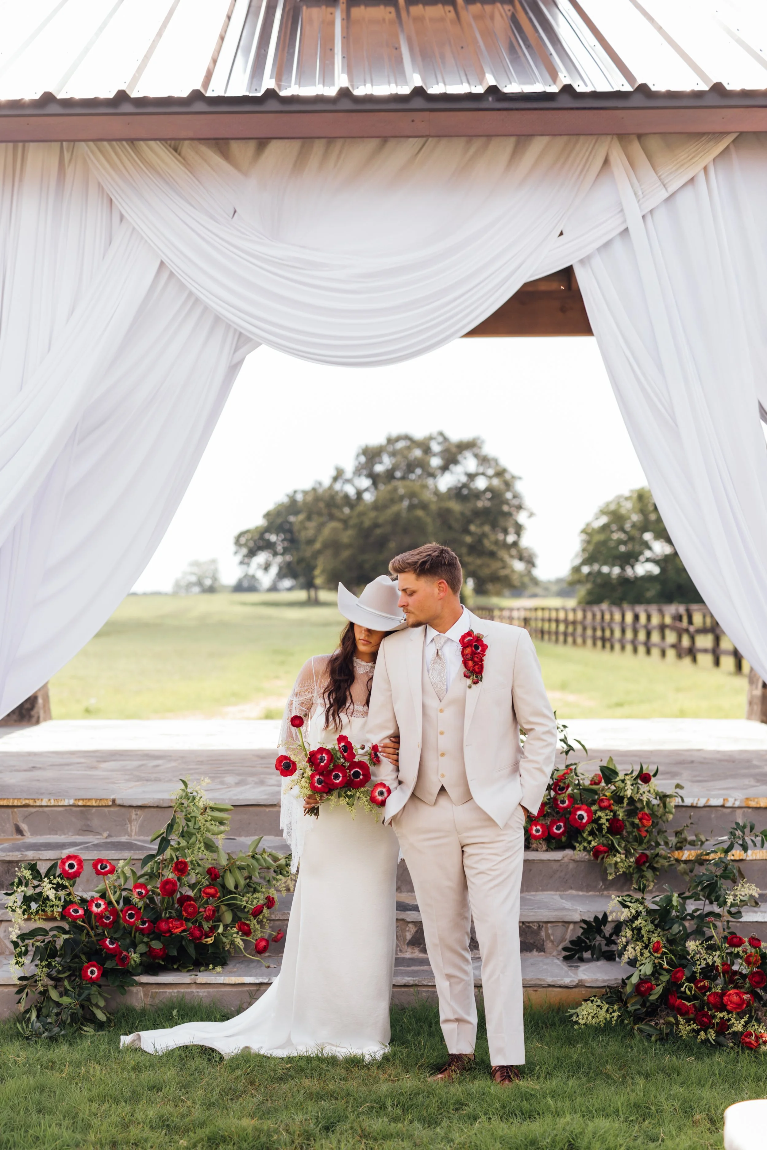 A newlywed couple stands under a white draped wedding arch decorated with red and black flowers, outdoors in a green field with trees in the background. The bride wears a white dress and veil, holding a bouquet of red flowers. The groom wears a light