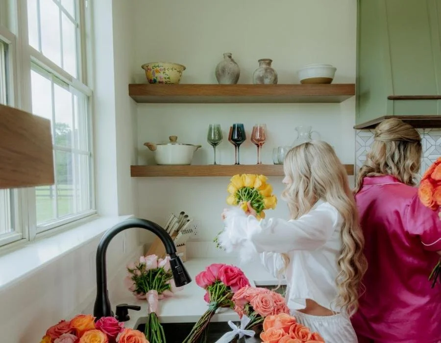 Two women with long, wavy blonde hair arranging floral bouquets in a bright kitchen with open shelves, colorful glassware, and a large window.