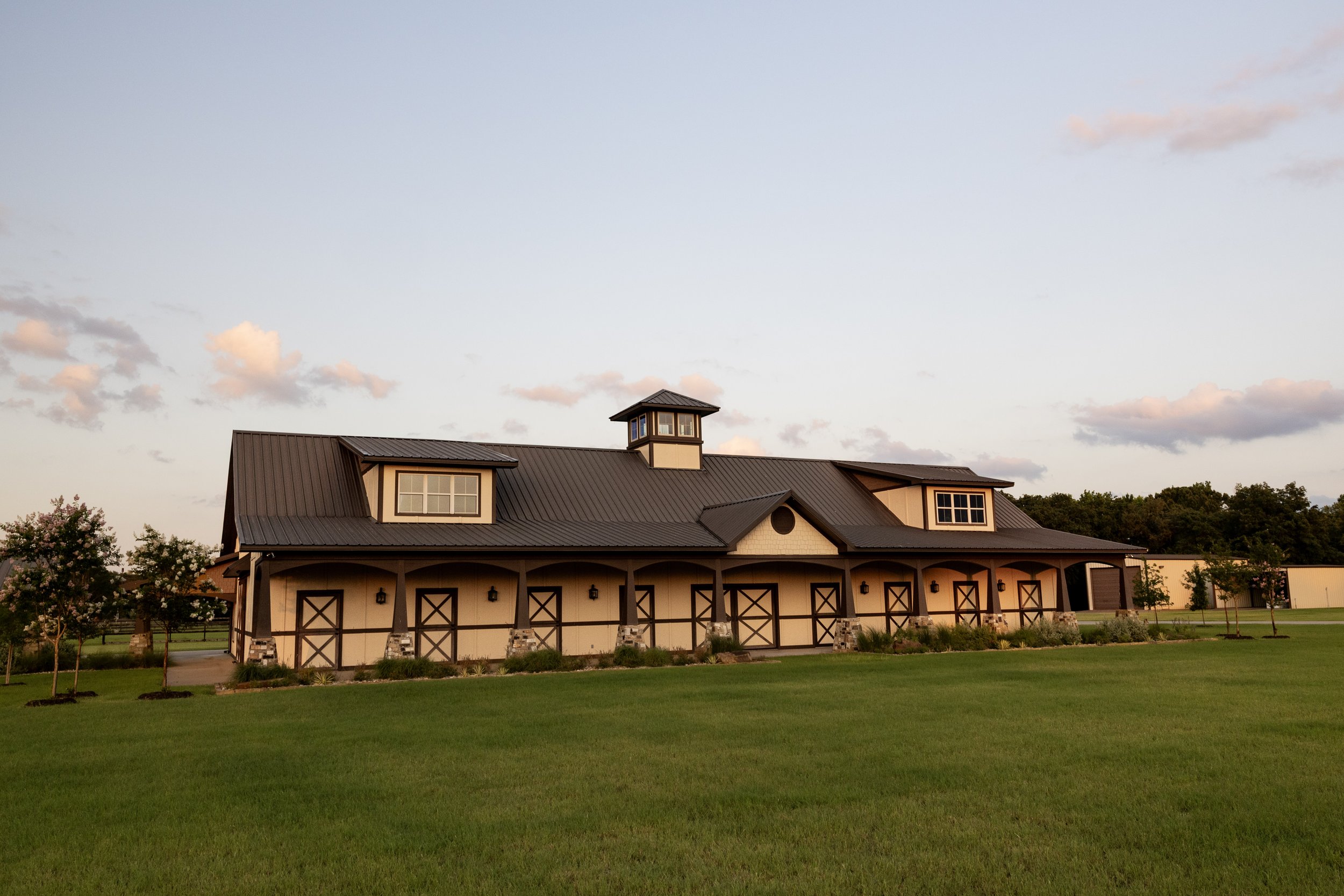 The stables wedding event space with a black metal roof, windmill cupola, and beige walls with dark brown accents, surrounded by green grass and trees, under a sky with light clouds during sunset.