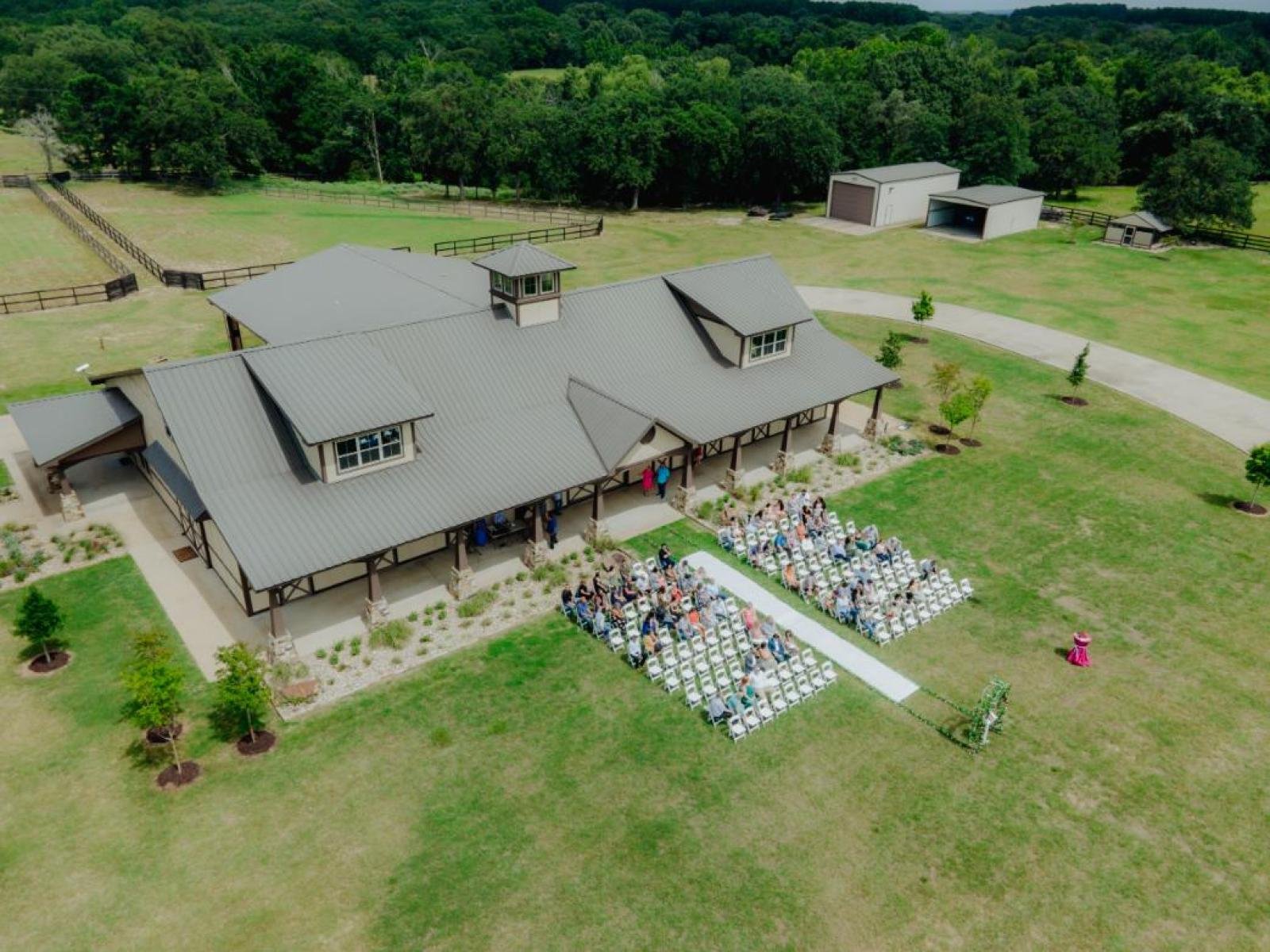 Aerial view of an outdoor wedding ceremony setup at a rustic building with a gray metal roof, with guests seated in rows of white chairs on a grassy lawn, surrounded by trees and open land.