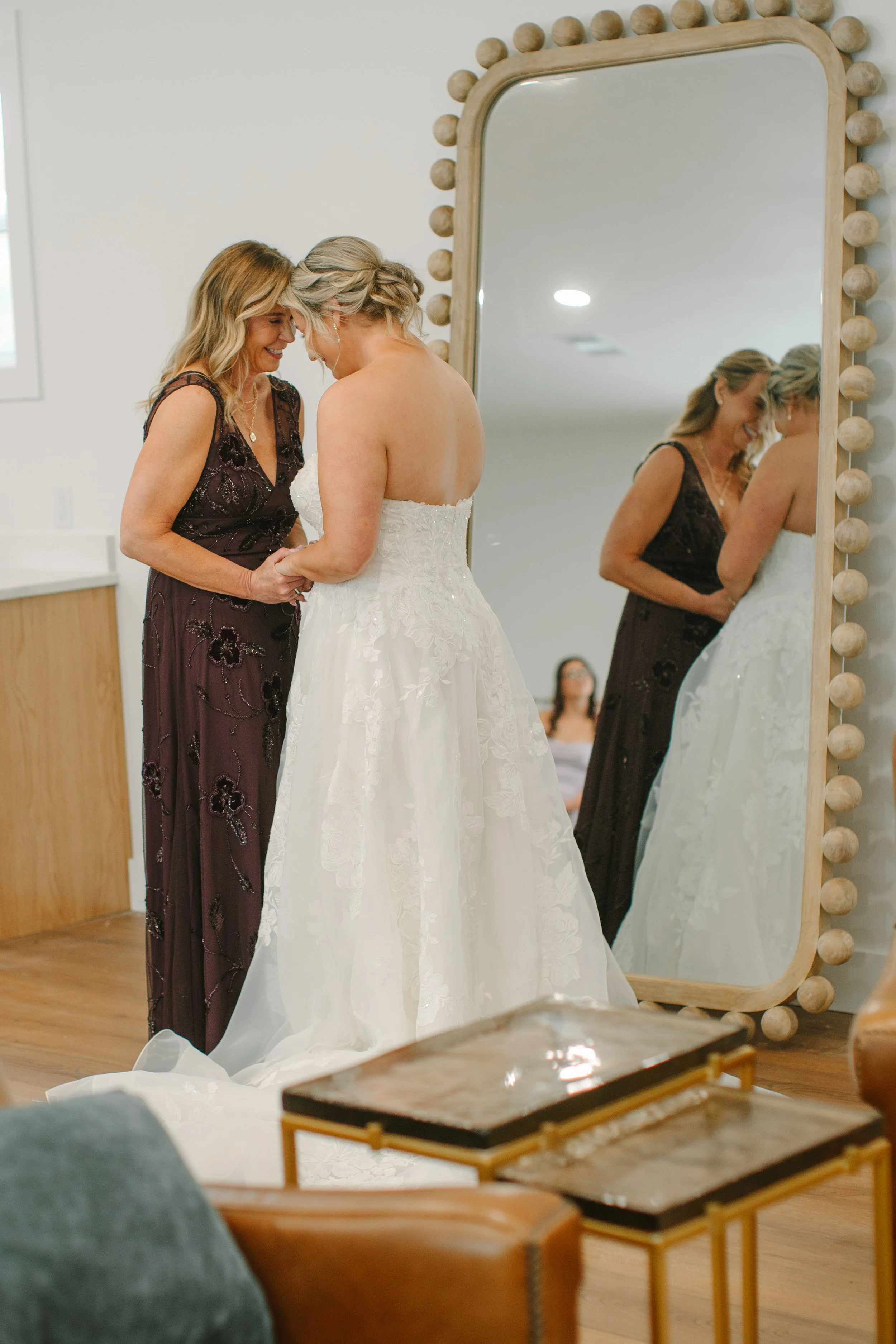 A woman in a strapless white wedding dress standing in front of a large mirror with decorative round light bulbs, adjusting her dress while smiling. An older woman in a dark, embroidered dress is helping her. The room has white walls and wooden floor
