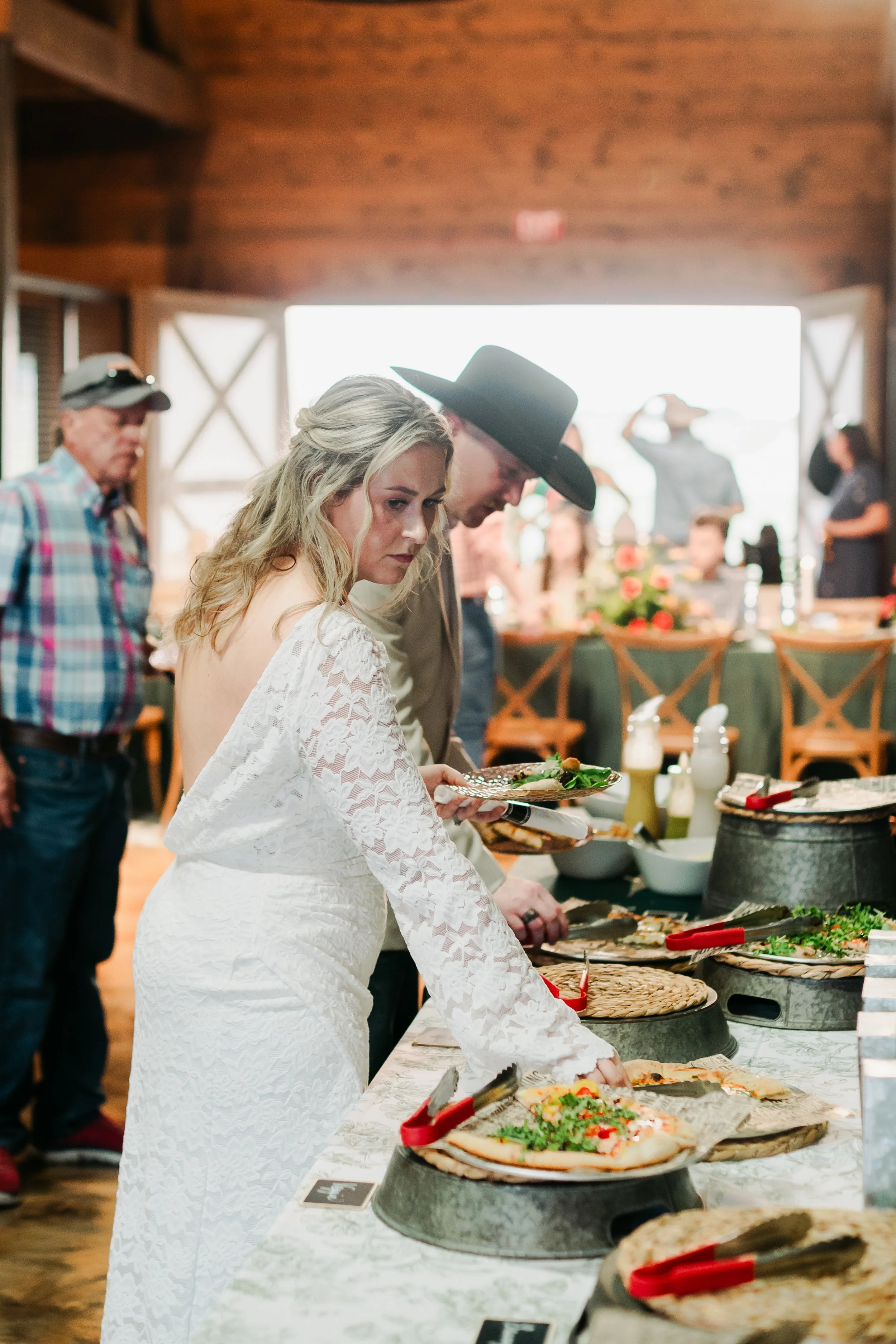 A woman in a white lace dress for a rehearsal dinner is selecting a pizza from a buffet table, surrounded by people in a rustic indoor setting with wooden walls and a large window in the background.