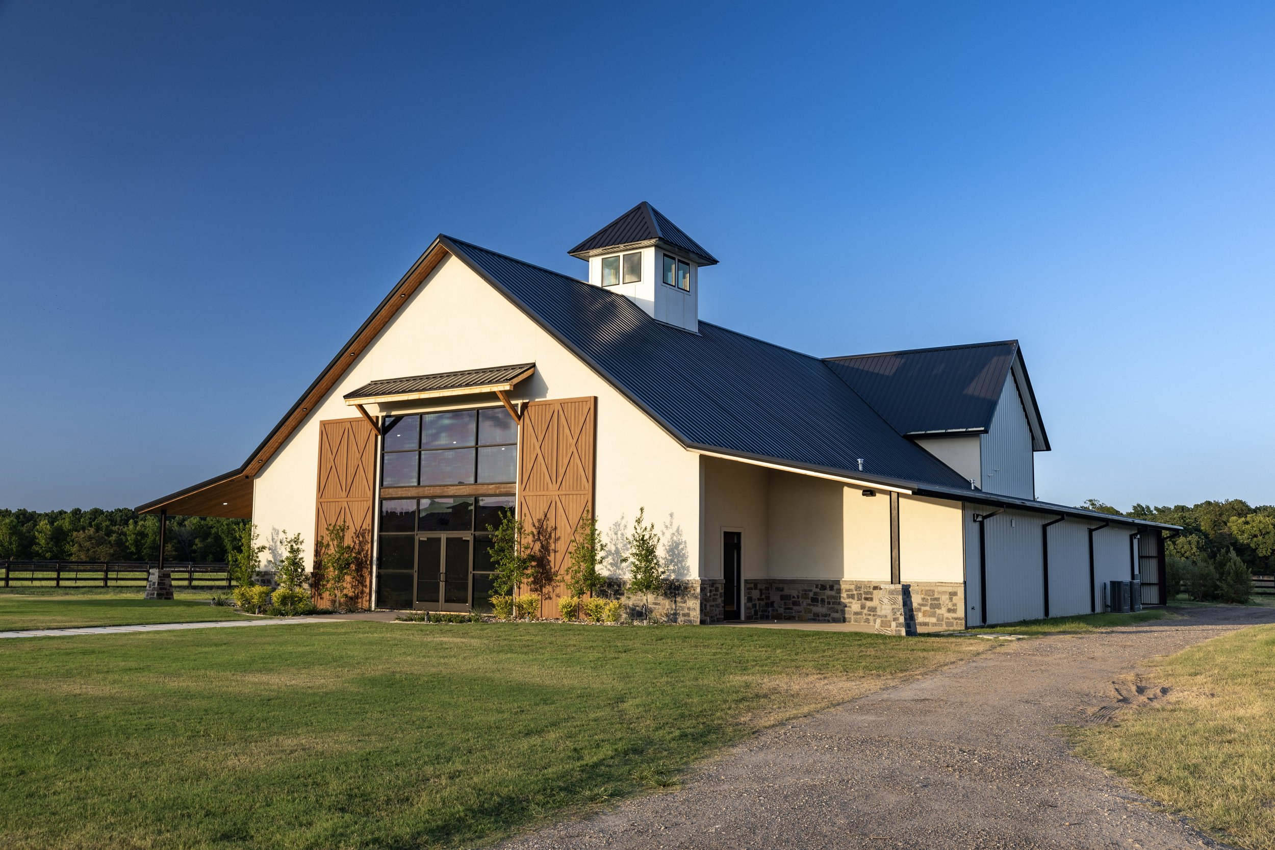 A modern house with a steep metal roof, large windows, wooden accents, surrounded by green grass and trees, under a clear blue sky.