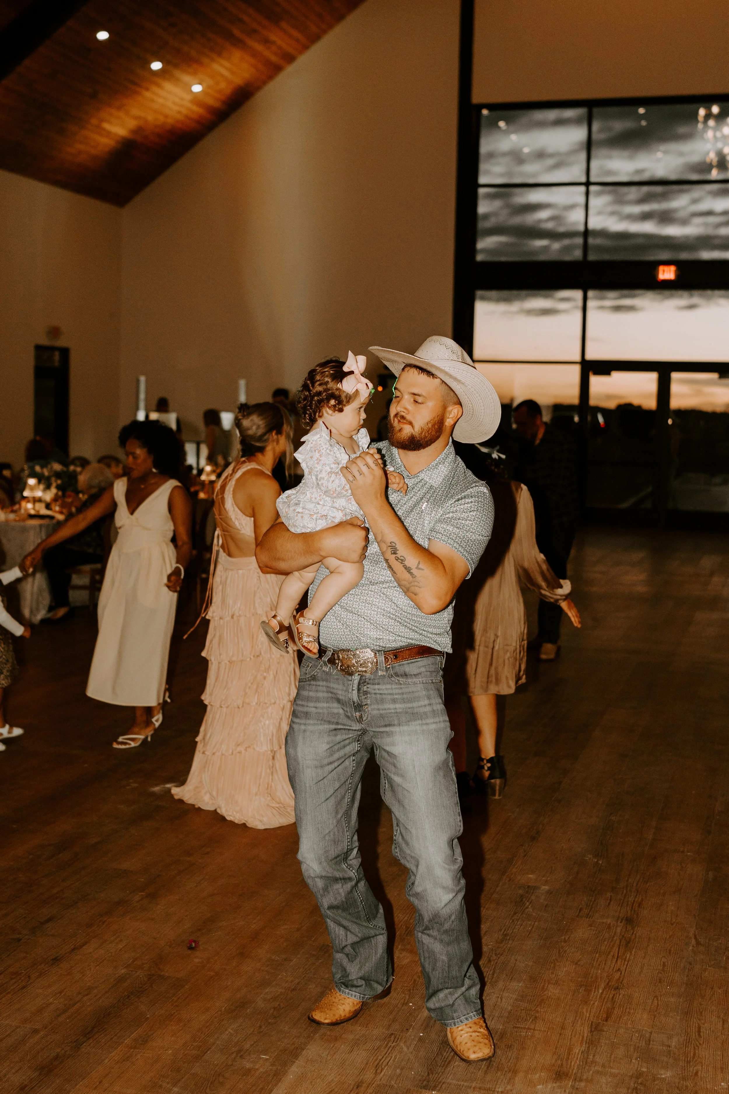 A man with a beard, wearing a cowboy hat, patterned shirt, jeans, and cowboy boots, is holding a young girl in a dress with a bow in her hair. They are dancing at an indoor event during sunset, with other people in the background.