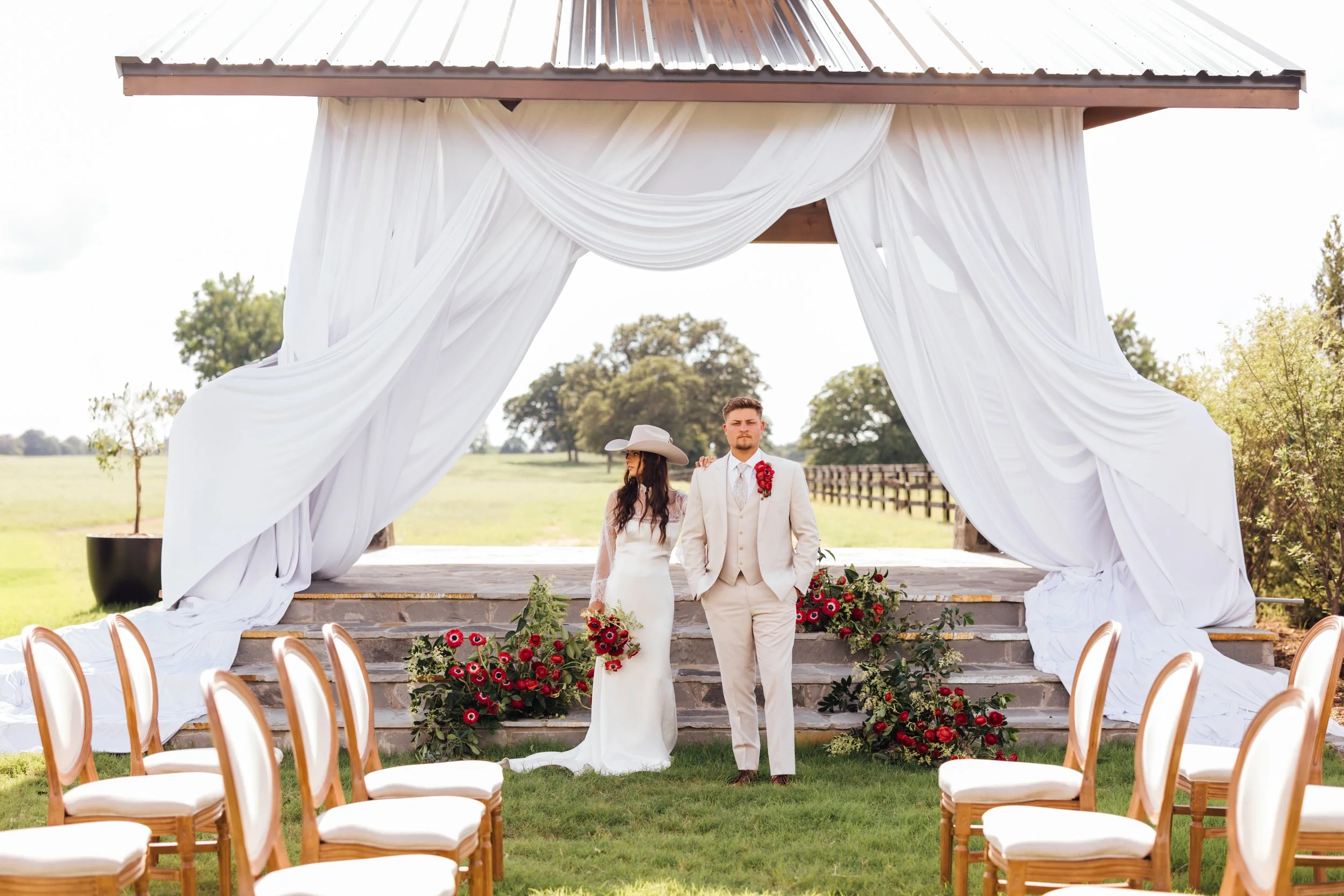 A couple getting married outdoors under a lux canopy with white drapes. The bride is in a white dress and the groom is in a beige suit with a red boutonniere. There are red flowers and chairs around them, and a grassy field extends in the background.