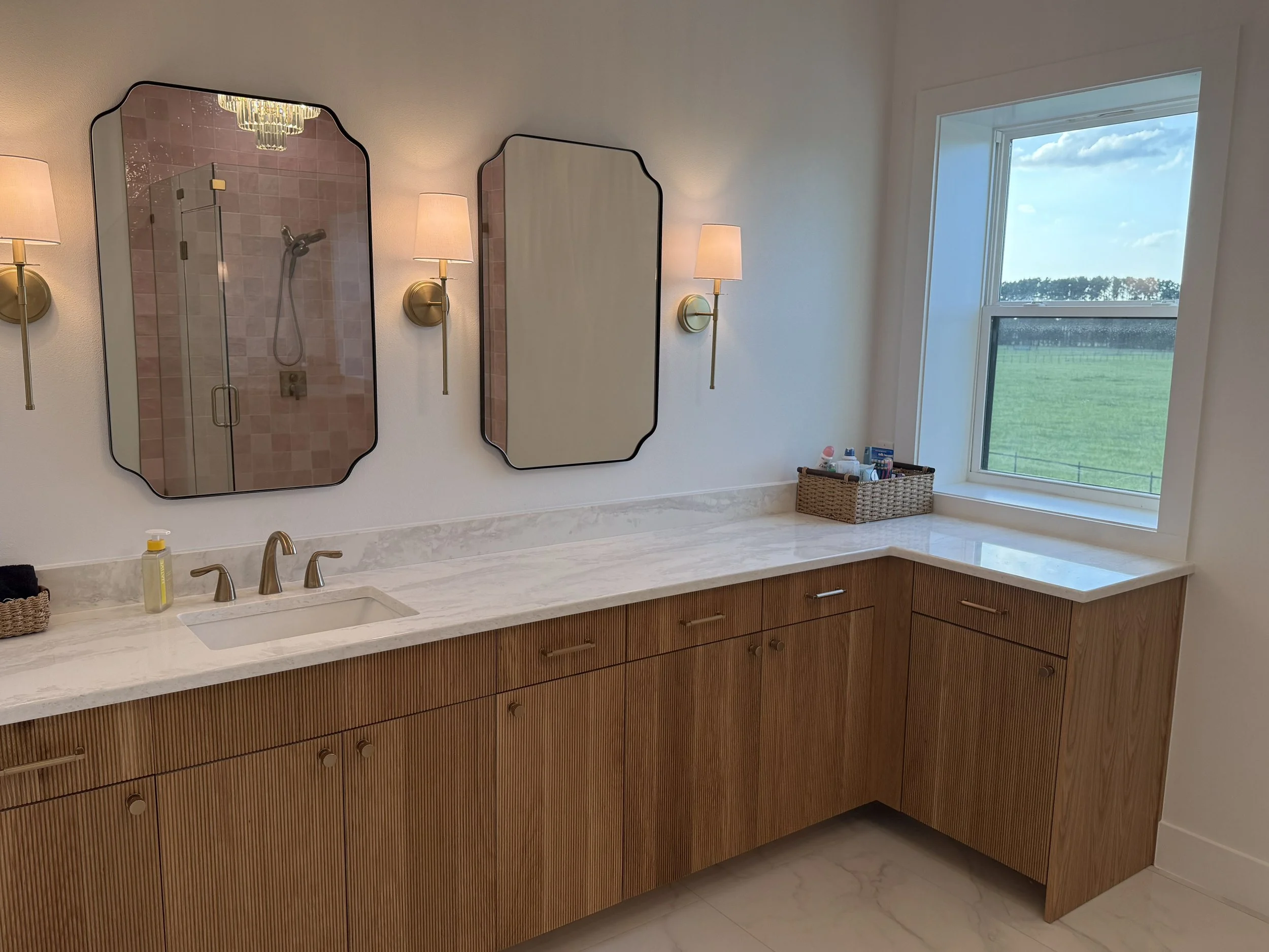 Bathroom vanity with marble countertop, wooden cabinets, two brass wall-mounted lamps with pink shades, two oval mirrors, and a window showing a green landscape with blue sky and clouds.