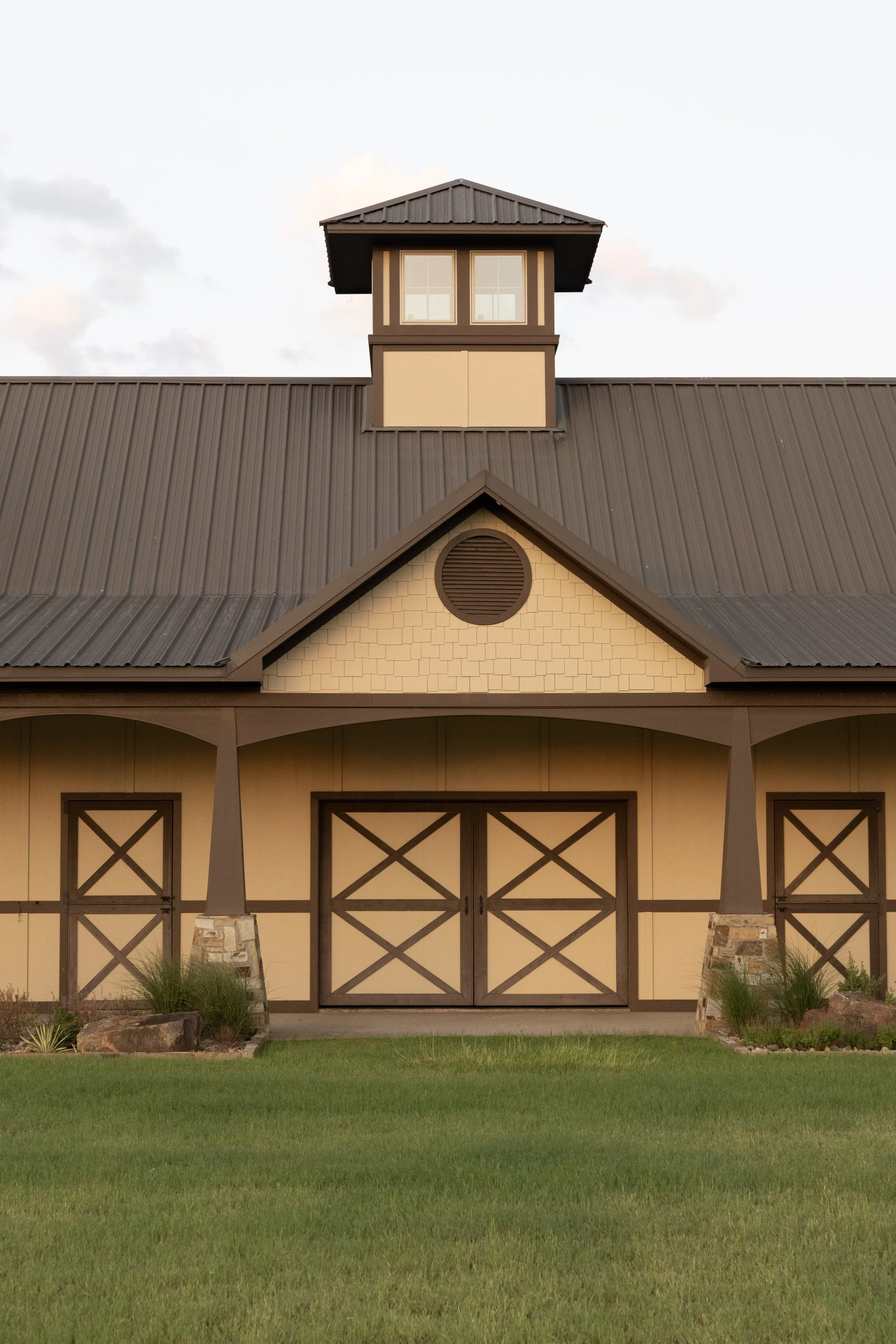 Front of an equestrian event space with a steep roof, decorative barn-style doors, a small windowed tower on top, and landscaped plants in the front yard.