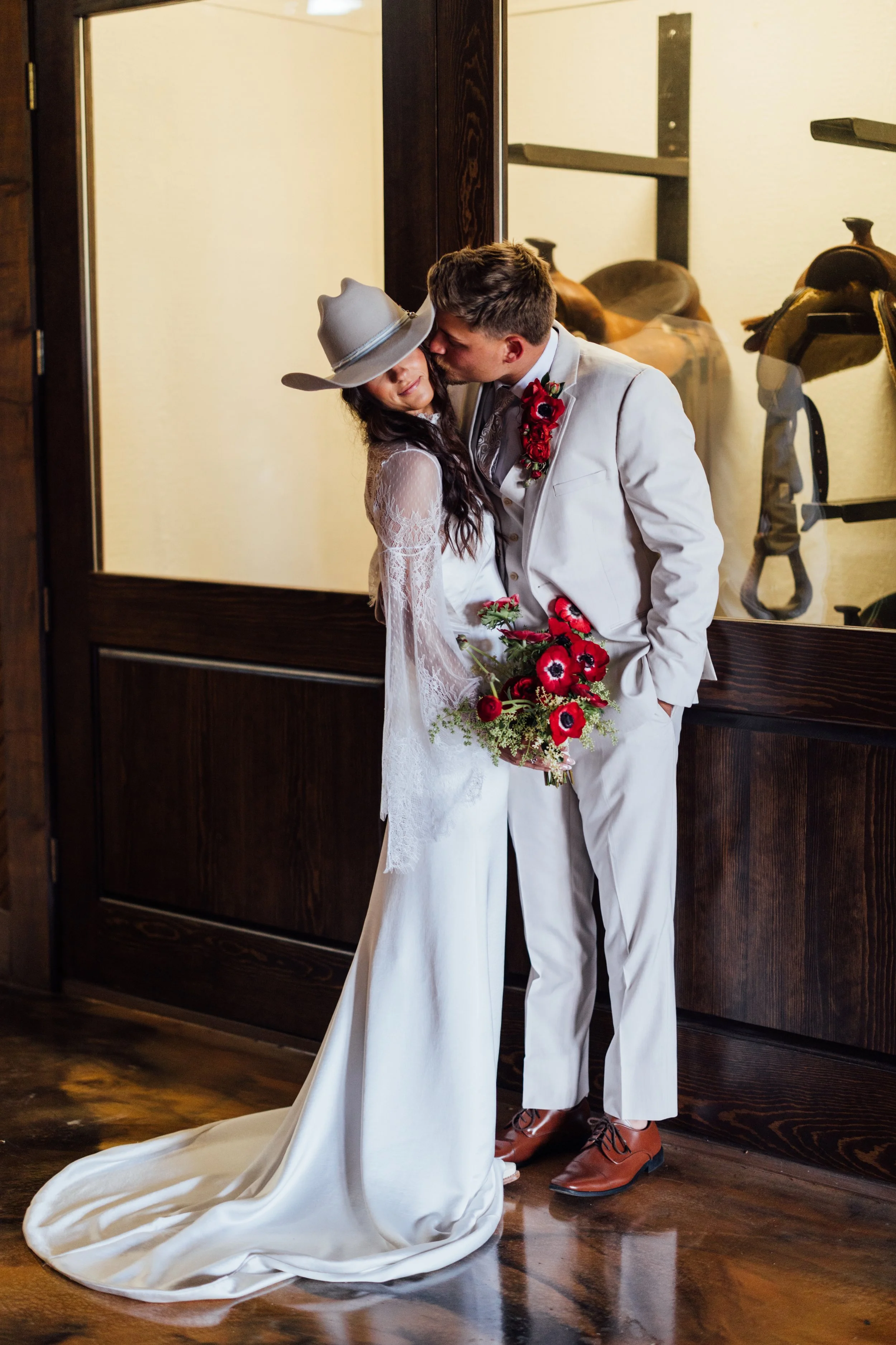 A couple dressed in wedding attire sharing a kiss, with the bride holding a bouquet of red and black flowers. The bride wears a white lace dress and a wide-brimmed hat, and the groom wears a white suit with a red flower boutonniere. They are standing indoors by a glass display case with saddles and riding gear visible.