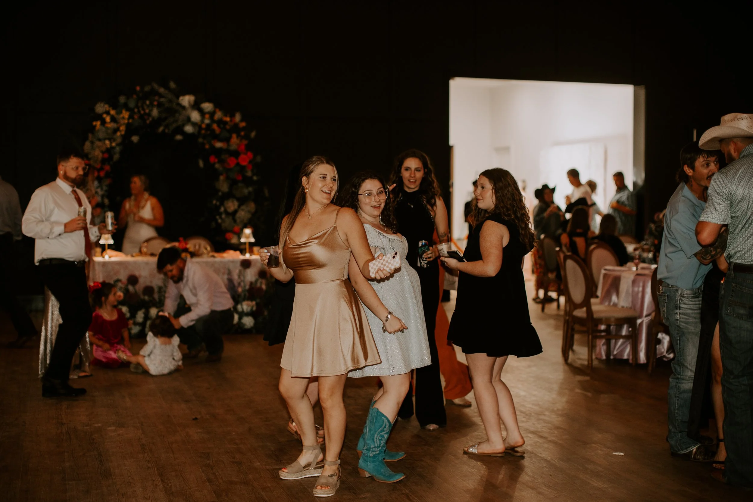 Four women dancing and talking at a social event in a decorated hall, with other guests and children in the background.