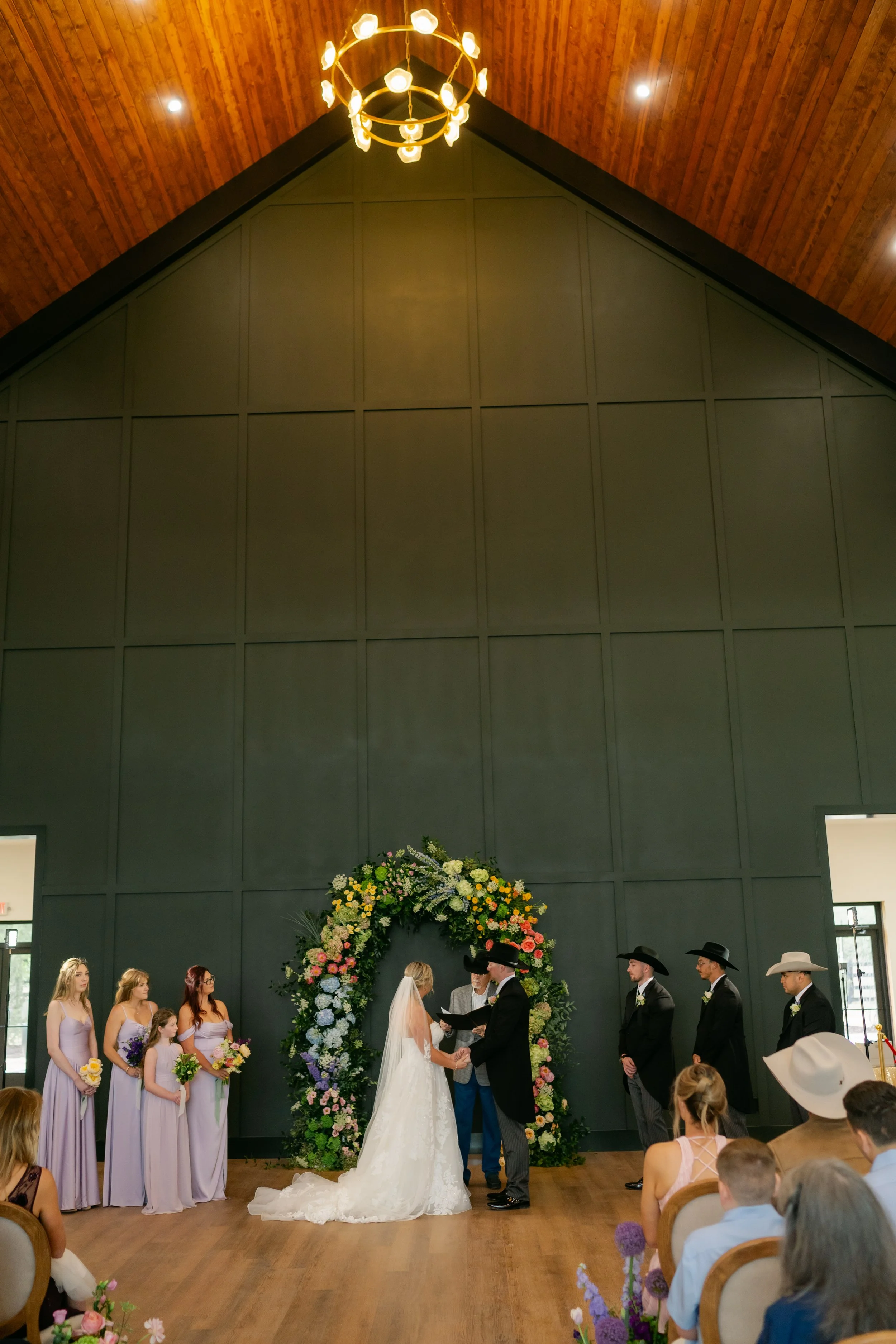 A wedding ceremony is taking place indoors in front of a dark green wall with a large floral arch. The bride and groom are exchanging vows, with the officiant in between them. The wedding party includes bridesmaids in lavender dresses holding bouquet