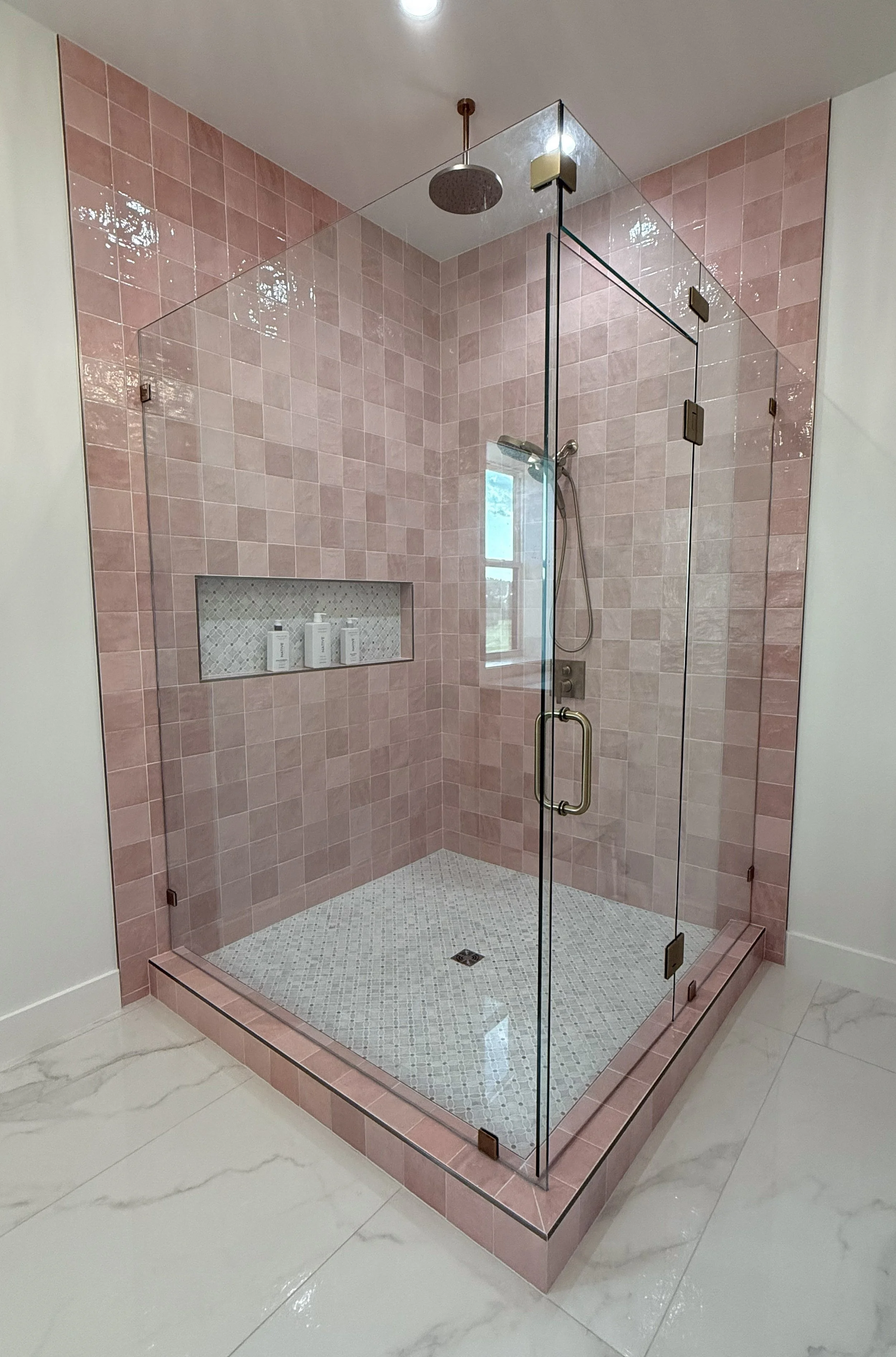 A modern walk-in shower with pink tiles, a glass enclosure, a rain showerhead at the ceiling, a handheld shower, a built-in niche with toiletries, and a white tiled floor, in a bathroom with marble flooring.