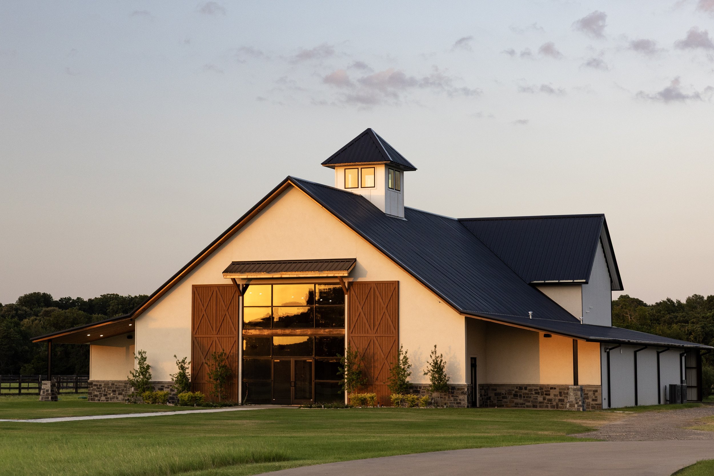 A modern farmhouse with a large front window, wooden accents, and a peaked roof, illuminated by sunset light, surrounded by green grass and trees.