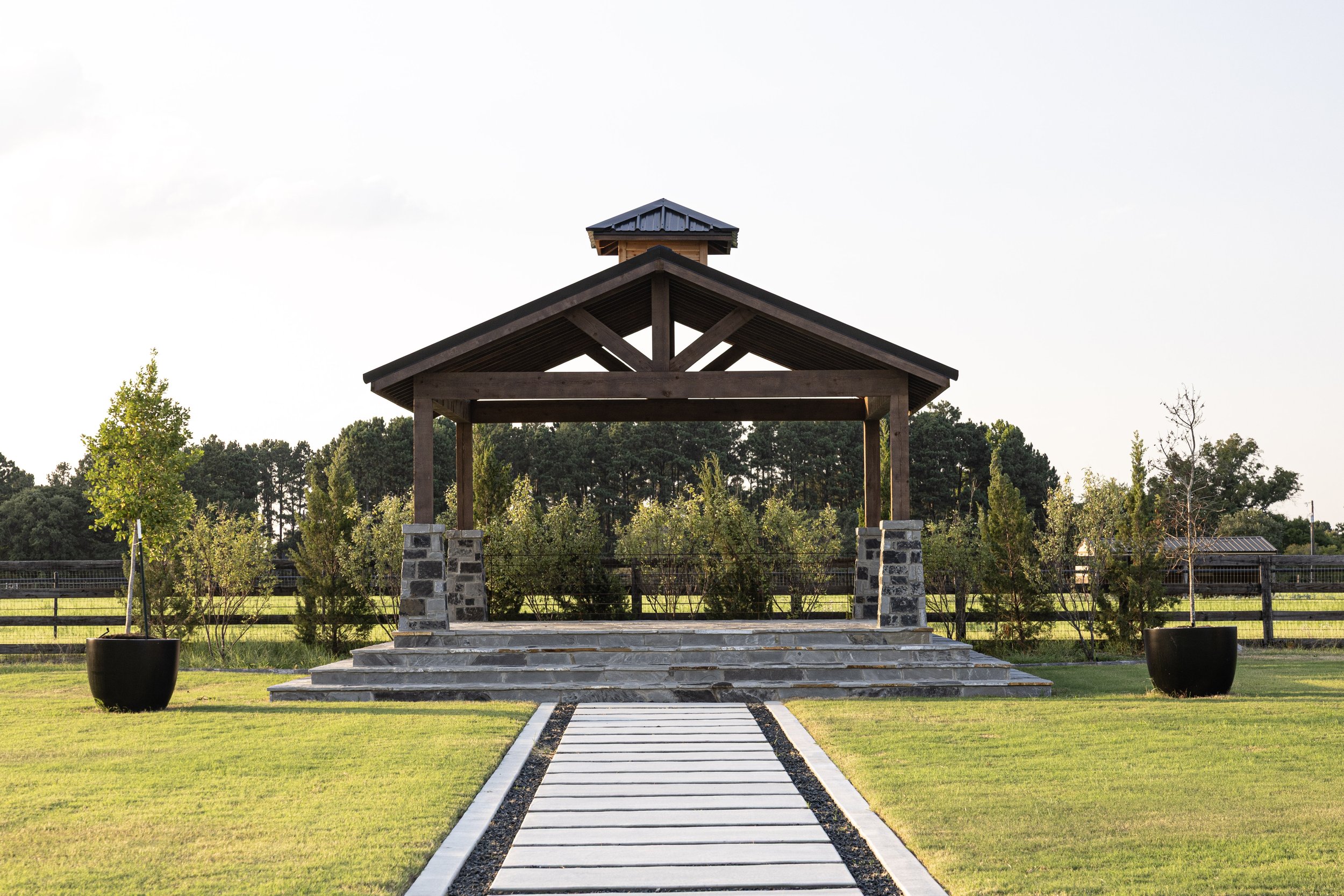 A wedding arbor landscaped outdoor area with a stone pathway leading to a wooden pavilion with steps, surrounded by green grass, potted plants, and trees, with a background of a fence and dense trees under a clear sky.