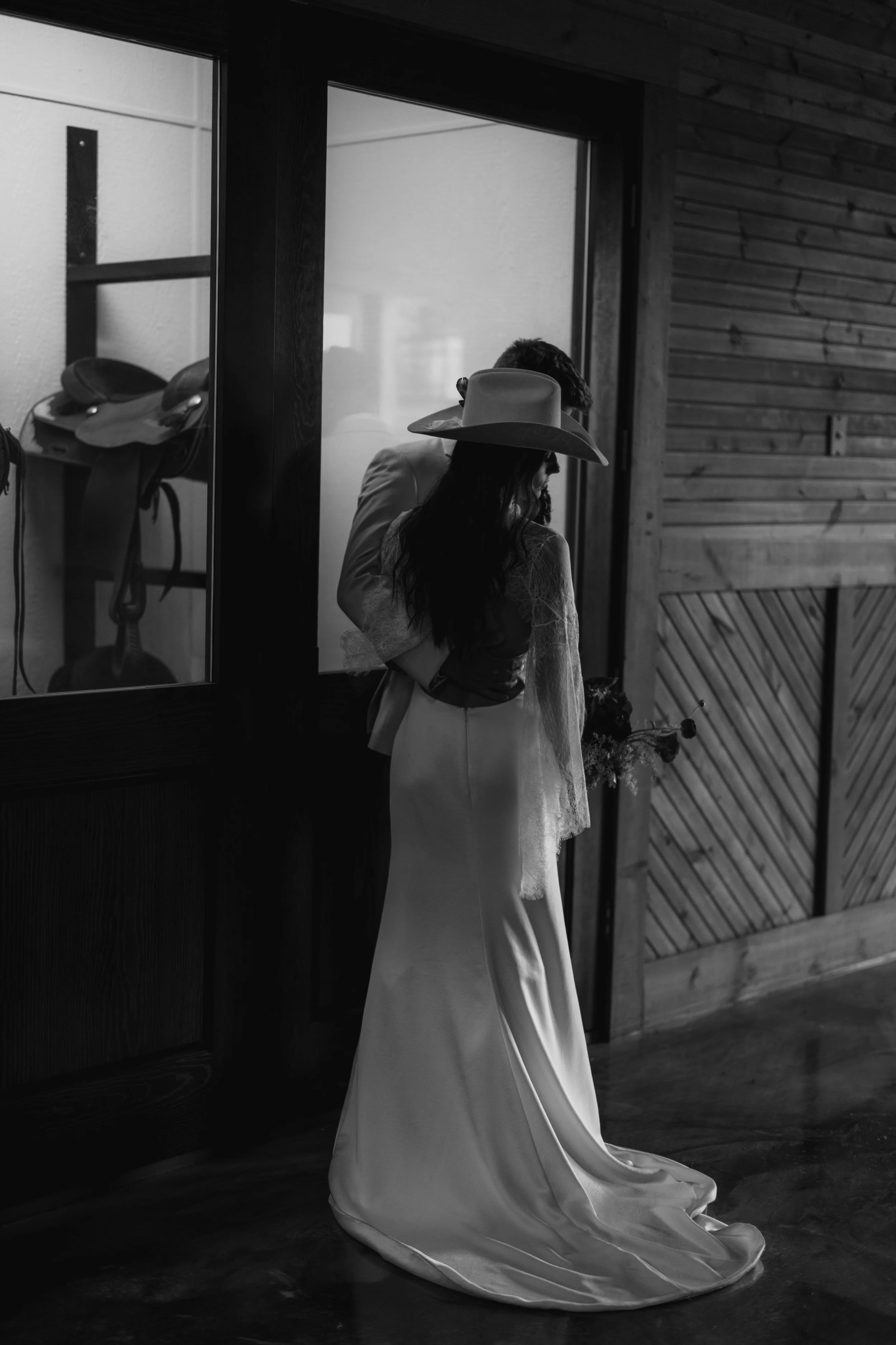 A woman in a long dress and wide-brimmed hat standing indoors near a wooden wall with another person partially visible behind her, holding a bouquet of flowers.
