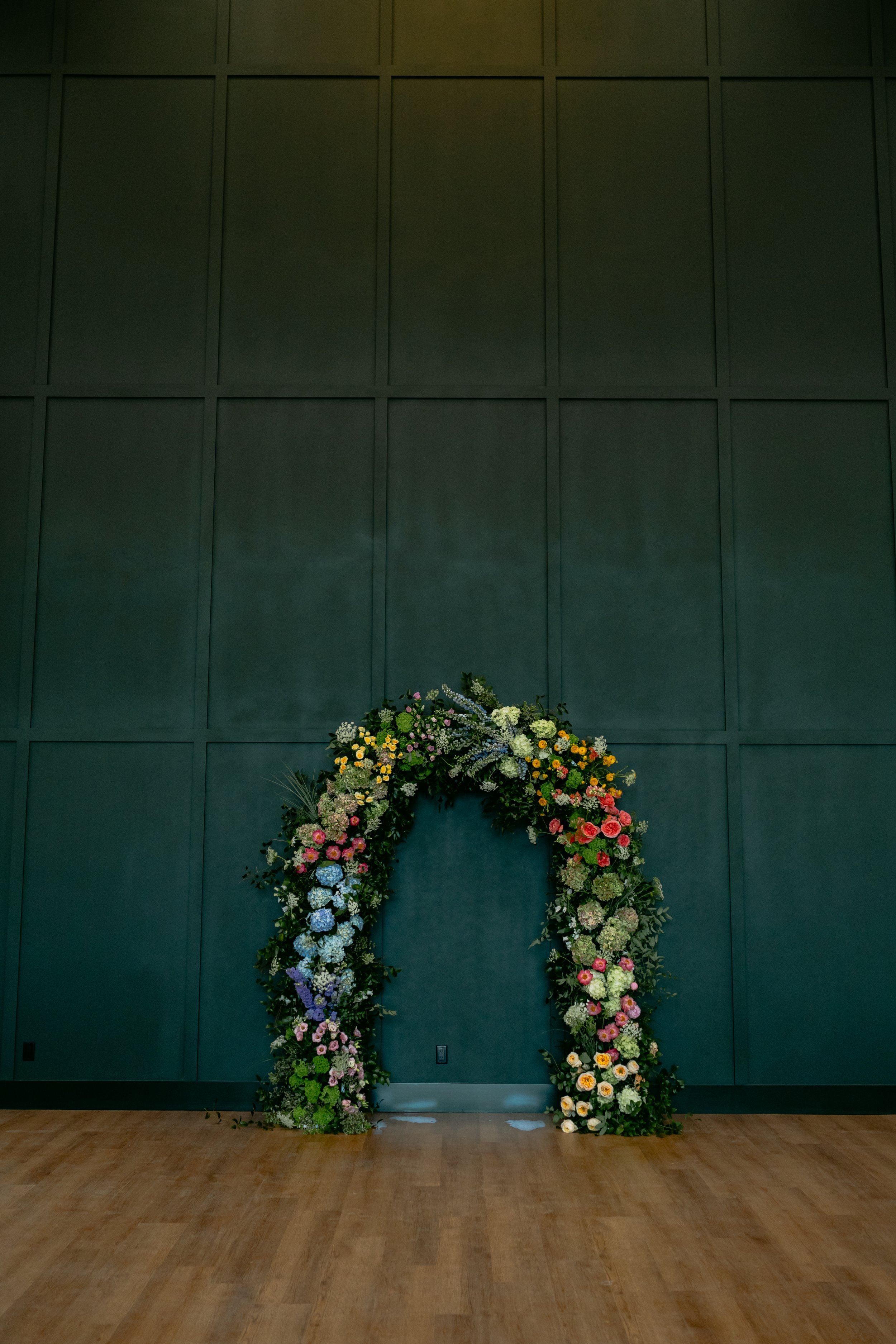 A floral arch made of colorful flowers is set against a dark teal wall with a wooden floor.