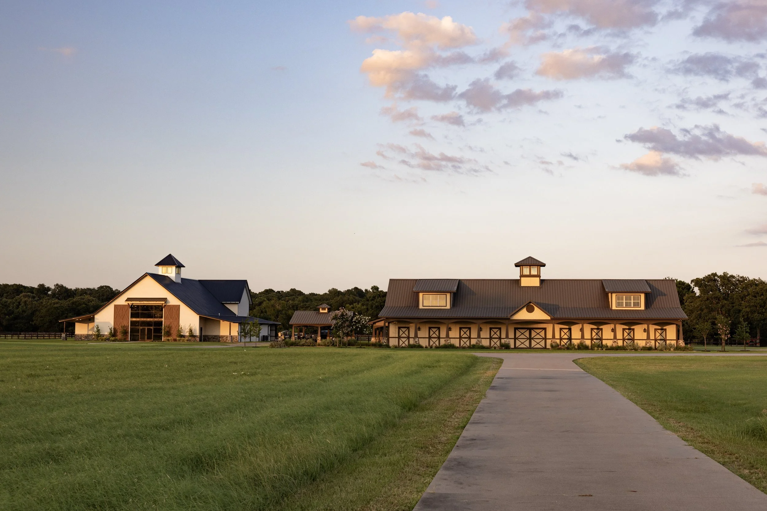A wide shot of a Indoor and Outdoor Wedding Venue for Luxury Weddings that hold up to 300 guests, manicured lawn, and a concrete pathway leading towards the buildings, with trees and a colorful sky with pink and blue clouds in the background.