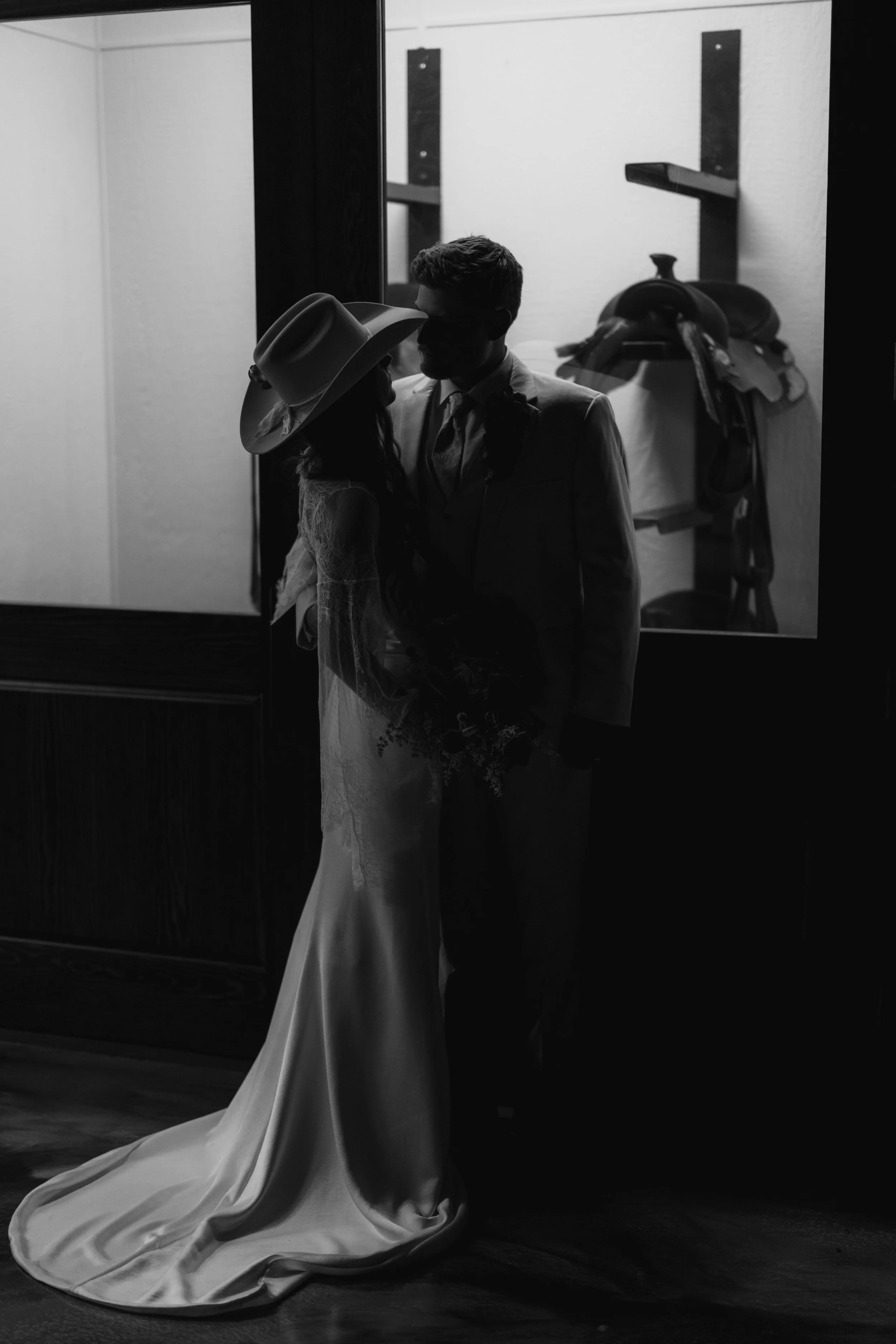 A black and white photo of a man and woman in wedding attire, standing close together, facing each other, with a mirror and a shelf with clothing items in the background.
