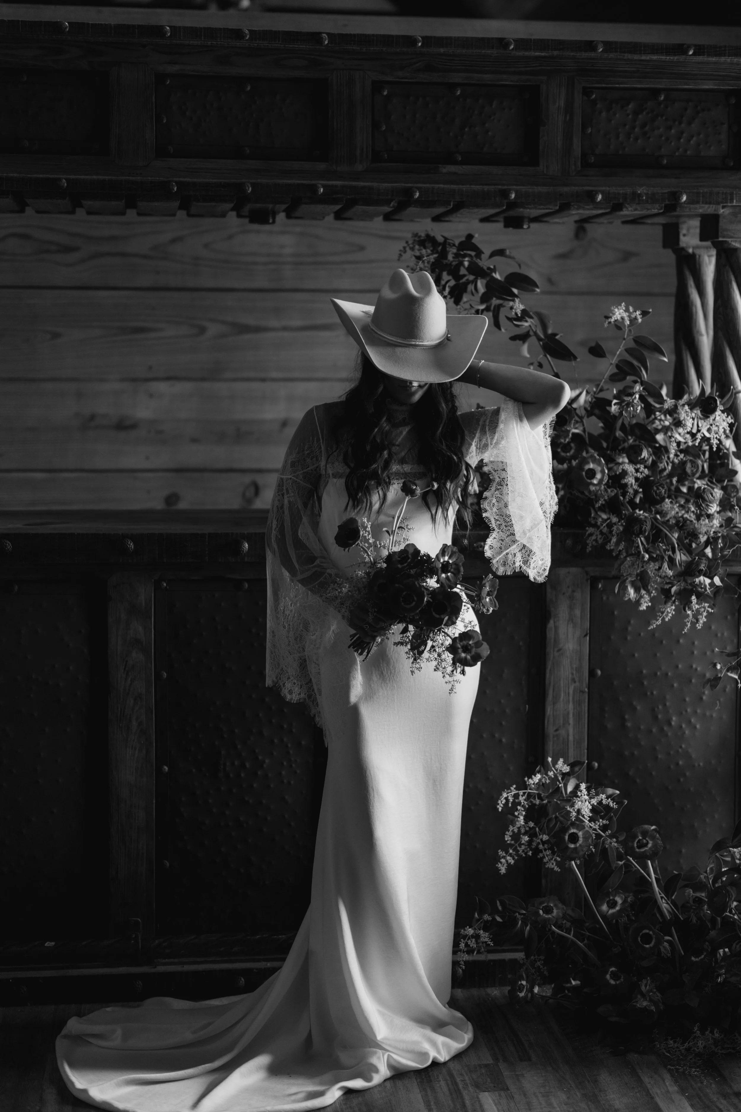 A bride wearing a large cowboy hat and a wedding dress standing in front of a rustic wooden barn built bar top.