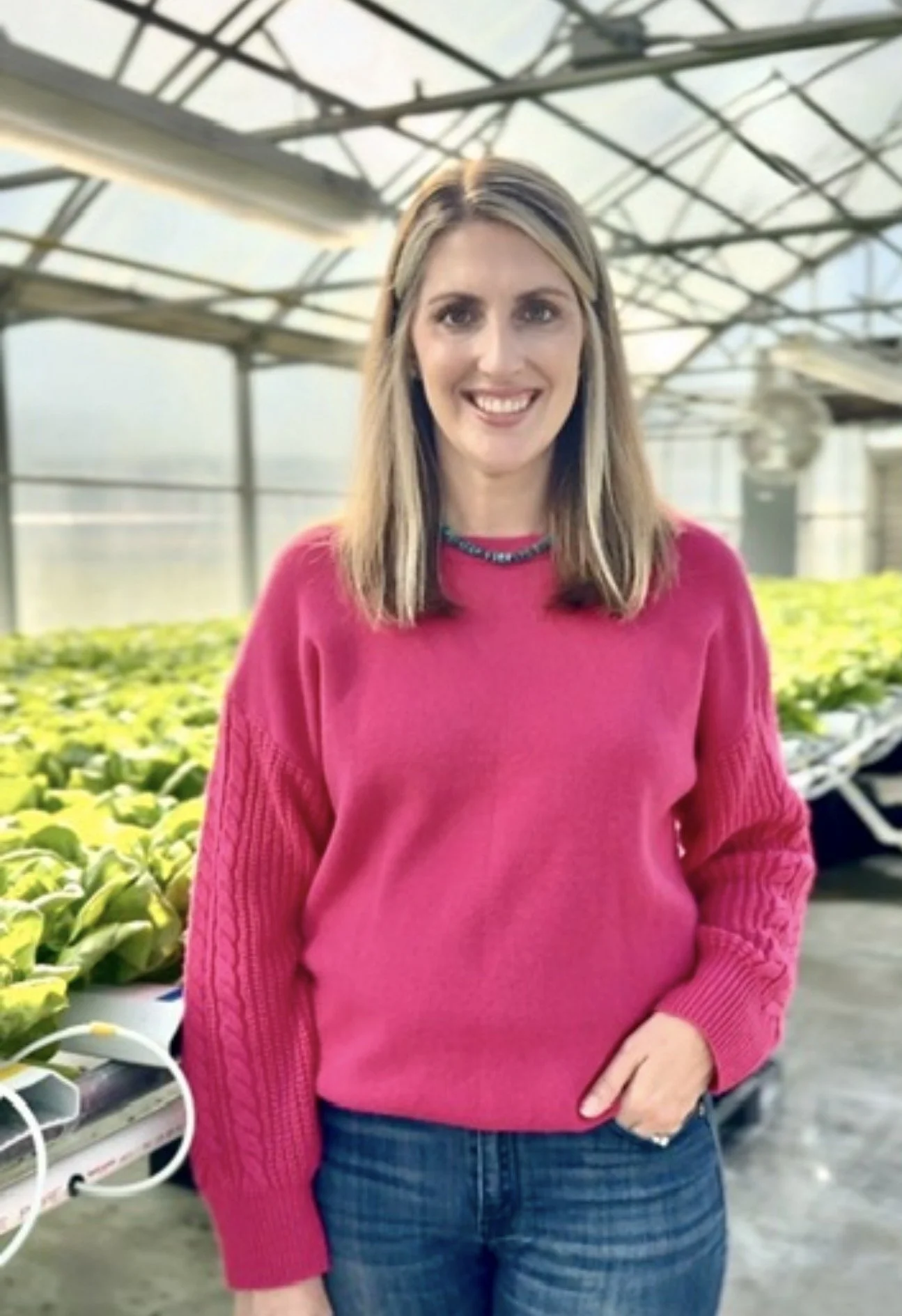 A woman in a pink sweater and jeans standing inside a greenhouse with plants behind her.