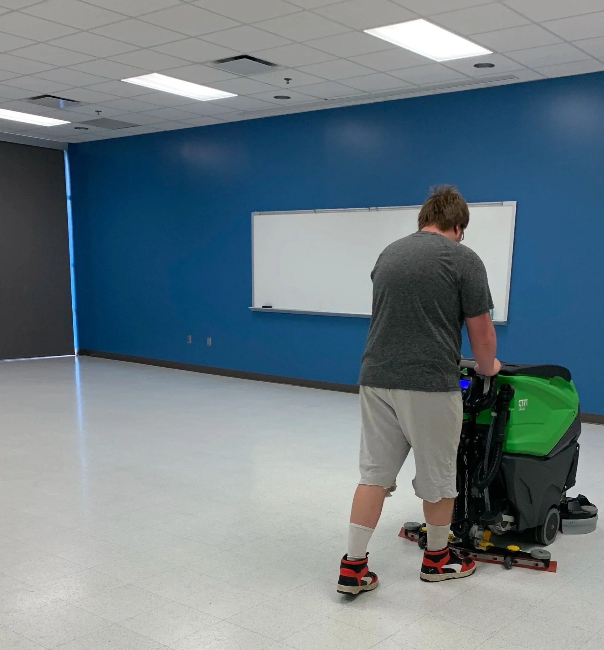 A man wearing a grey t-tshirt, gym shorts and sneakers, pushing a floor cleaning machine in an empty room with a whiteboard and blue walls.