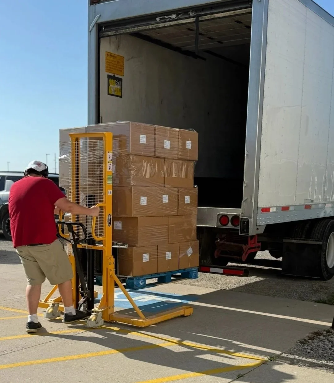 Person operating a pallet jack loaded with boxes, loading produce into a semi truck outside.