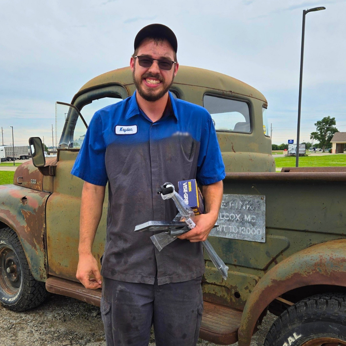 A man smiling and wearing a blue mechanic shirt stands in front of an old, vintage pickup truck outside. He is holding a small box of tools.