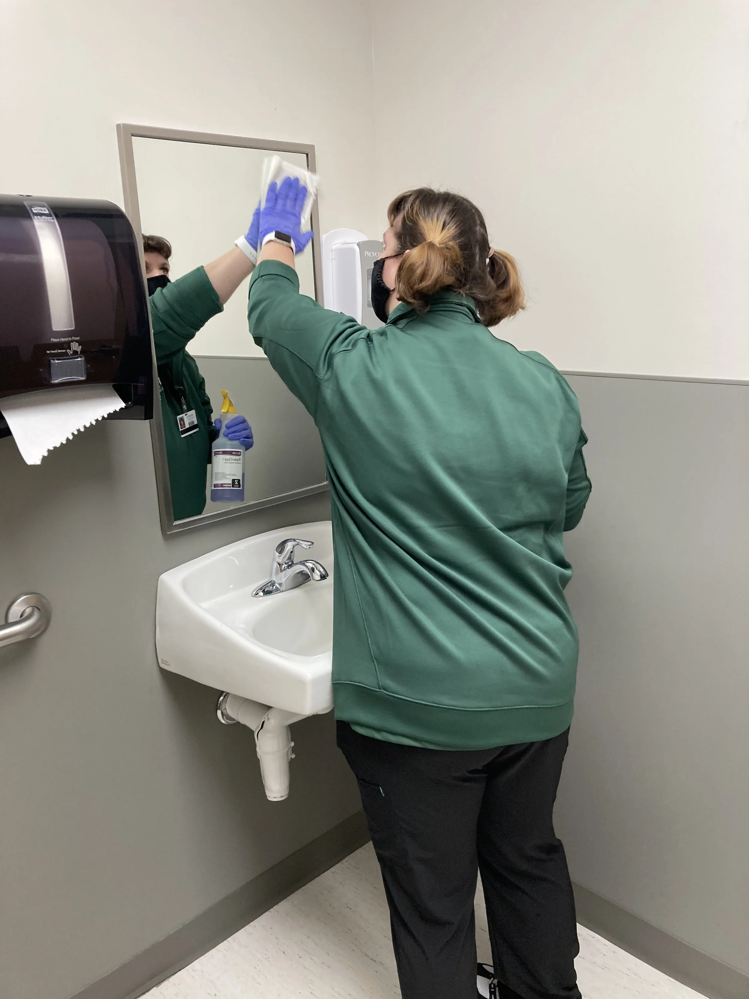 A healthcare worker disinfecting a bathroom mirror in a medical facility.