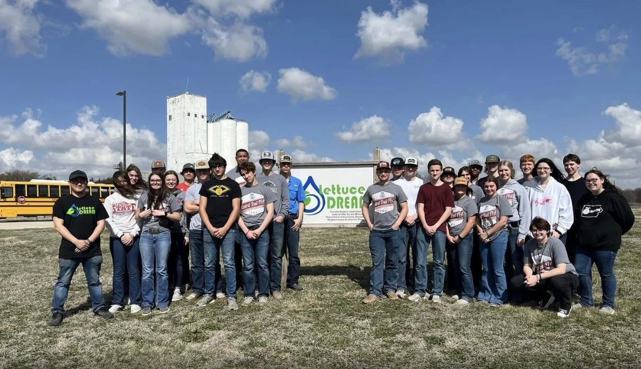Group of students standing on grass field in front of a sign that says Lettuce Dream, with a yellow school bus and farm silos in the background under a partly cloudy sky.