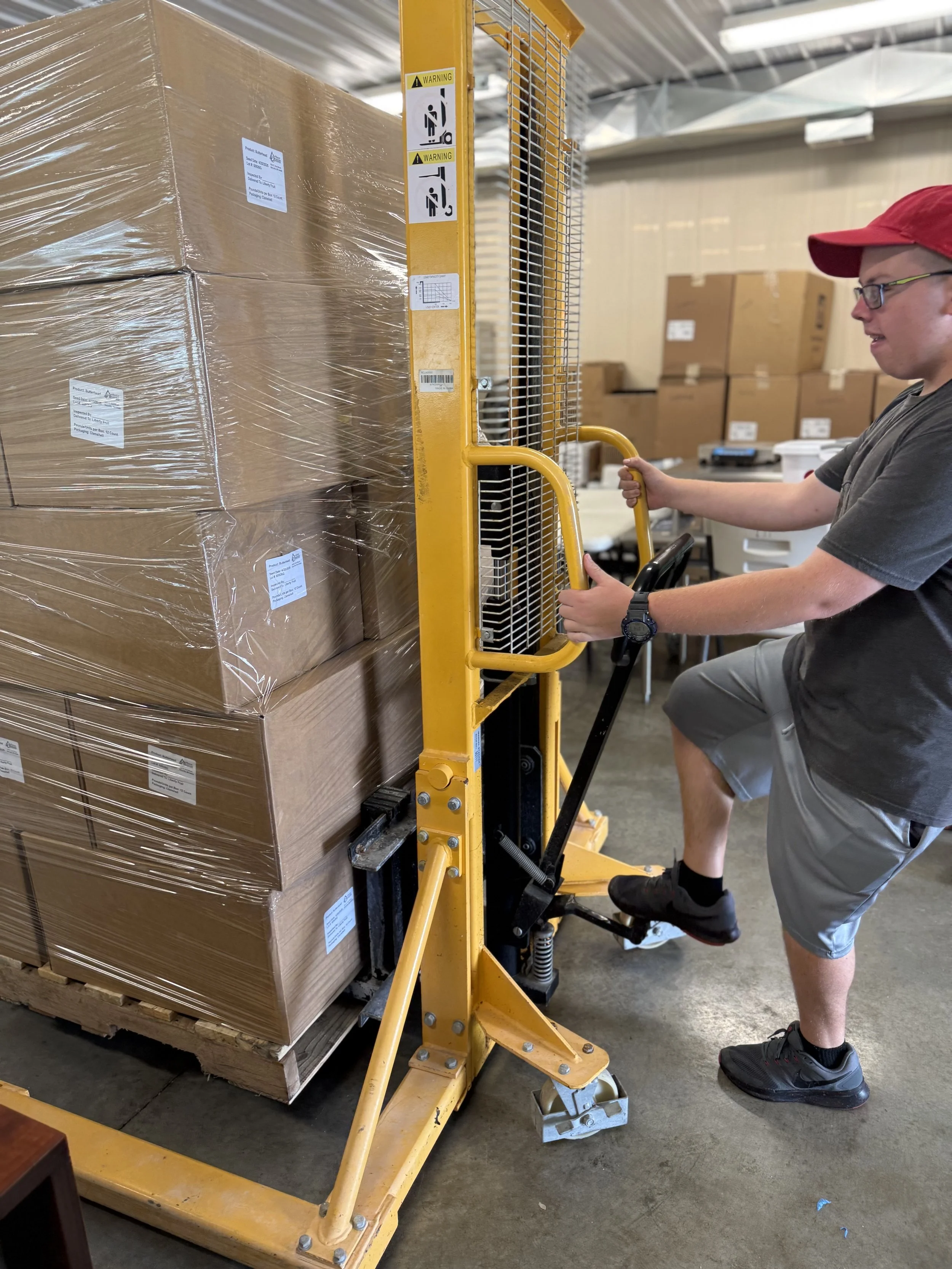 A young man adjusting a yellow hand-operated pallet lift with large pallets of boxes wrapped in plastic in an operations room.