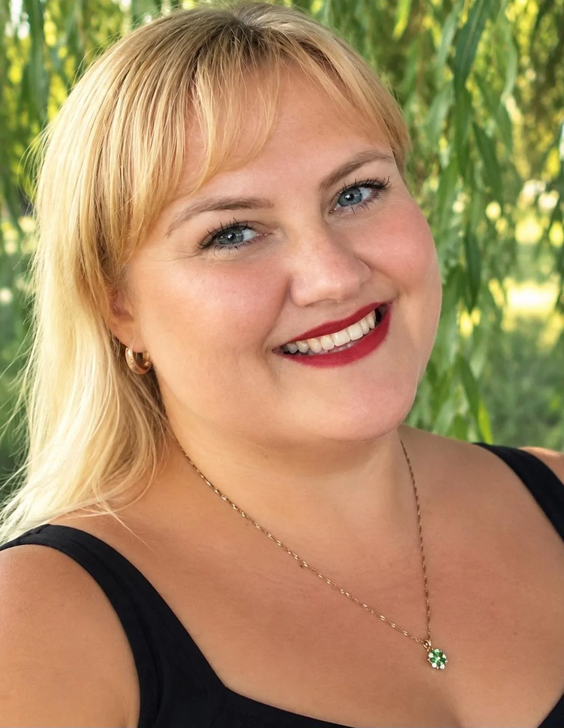 Nahaufnahme eines Lächelns einer Frau mit blonden Haaren, blauen Augen und rotem Lippenstift, vor grüner Natur im Hintergrund.