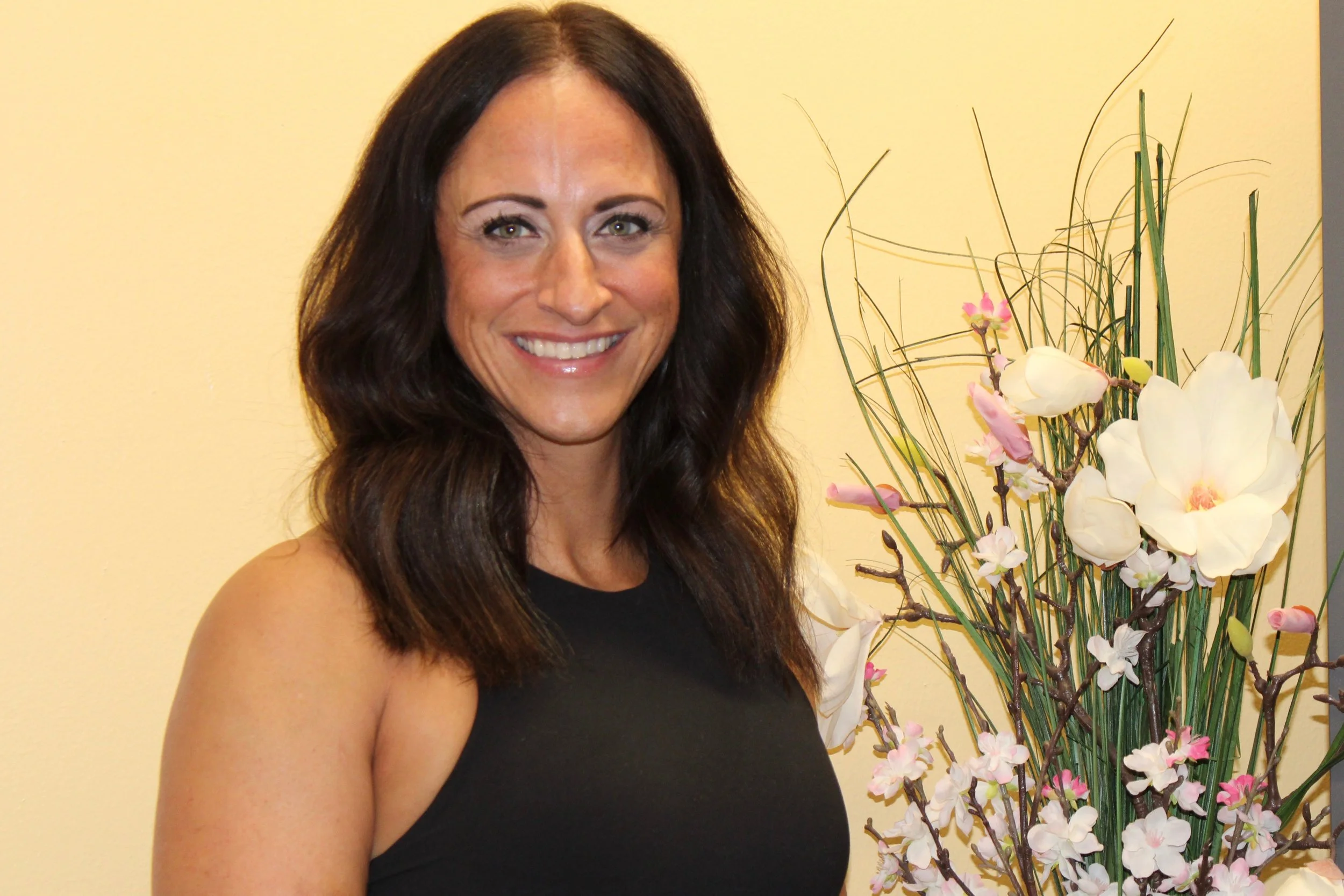 A woman with long dark hair smiling and standing next to a large arrangement of white and pink orchids and tall green foliage.