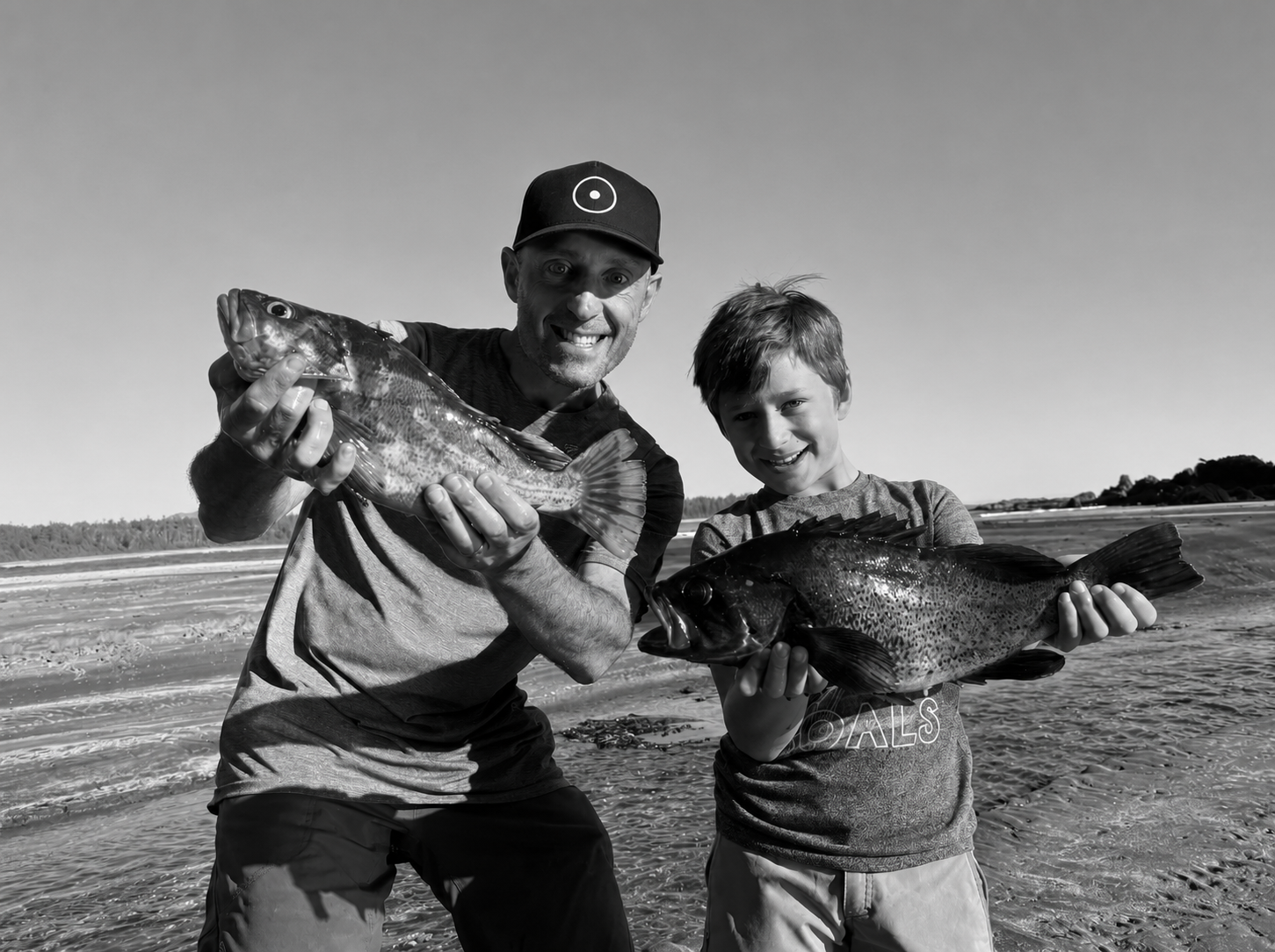 A man and a boy holding large fish on a sandy beach, smiling, with the ocean in the background.