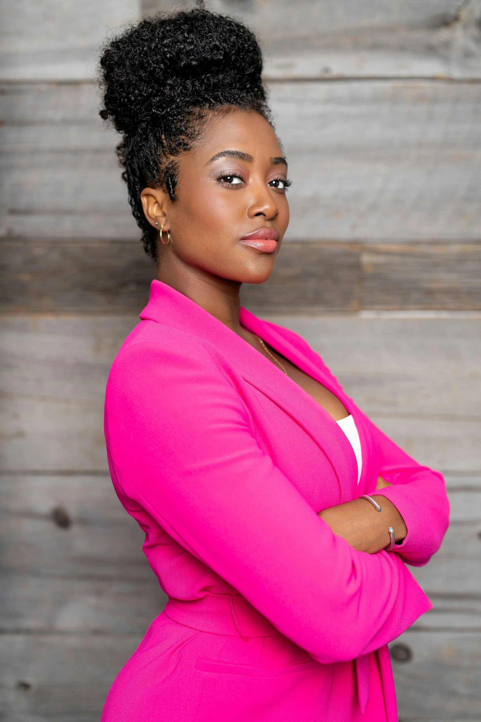 A woman with dark, curly hair styled in a high bun, wearing a bright pink blazer, standing with arms crossed in front of a wooden wall.