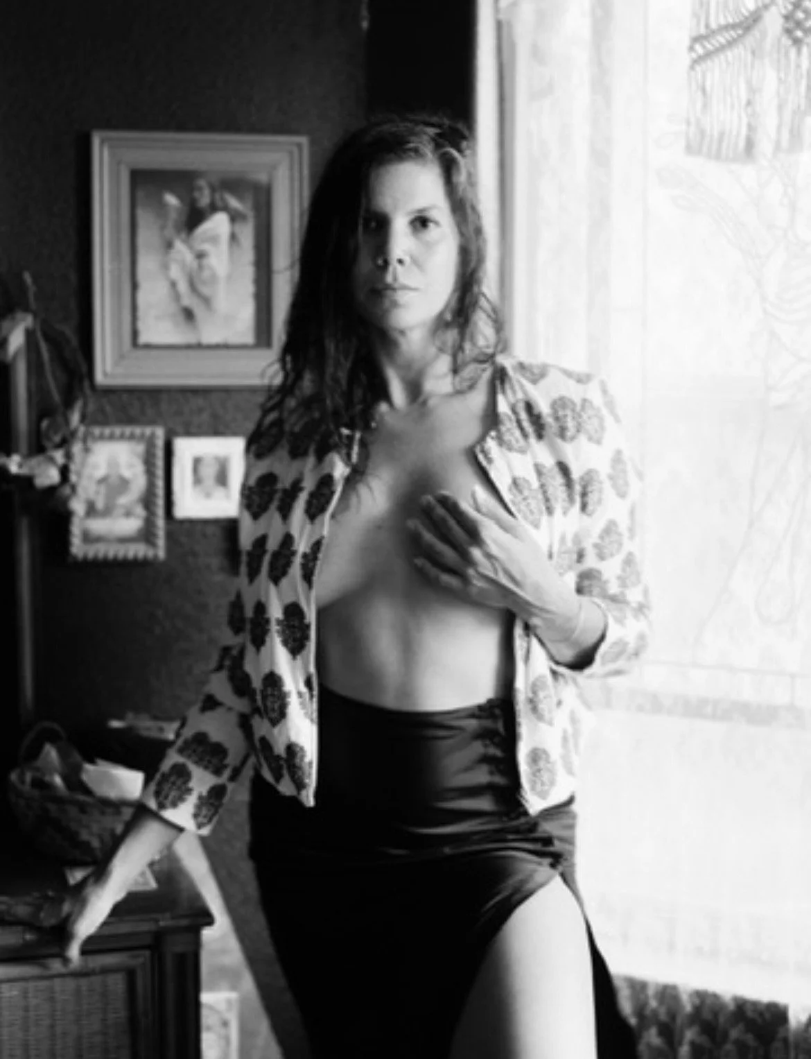 A black and white photo of a woman standing indoors near a window, holding her hand over her chest, wearing a patterned shirt and a skirt, with framed pictures and a painting on the wall behind her.