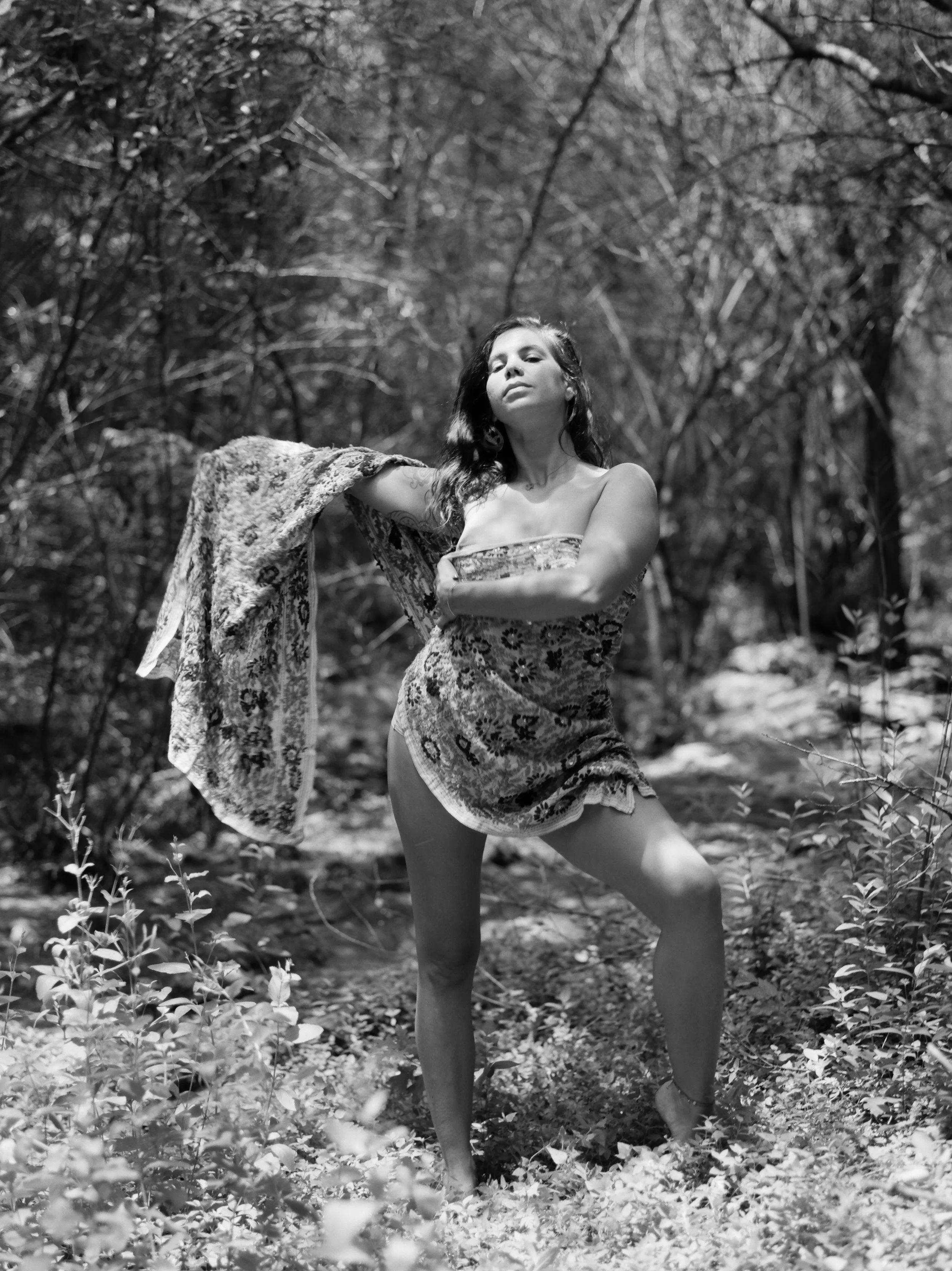 A woman with long dark hair, standing outdoors in a wooded area, wearing a patterned dress, holding a piece of fabric, with a confident expression and muscular stance.