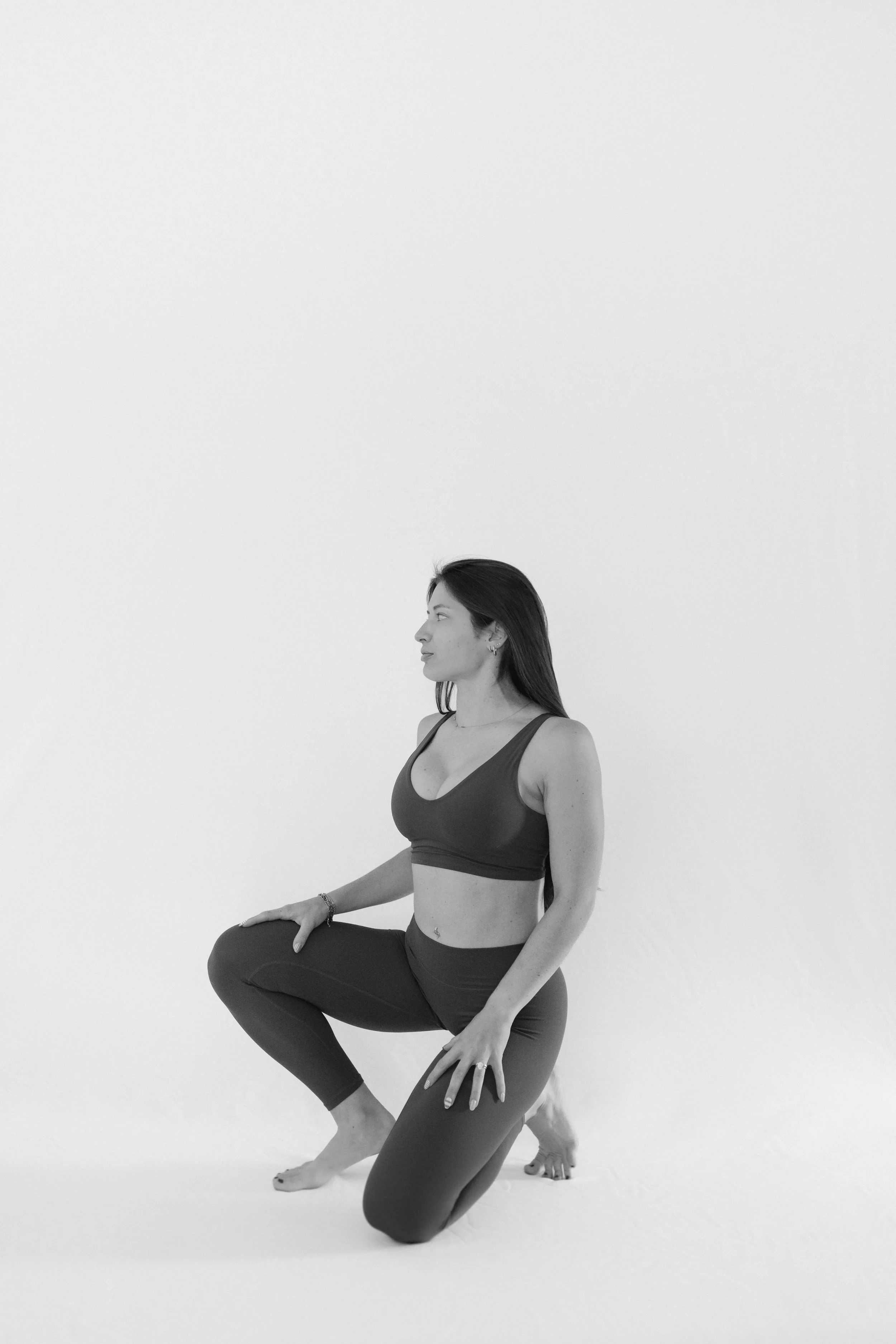A woman in athletic wear doing a yoga pose with one arm on her knee and the other arm relaxed, with her eyes open, against a plain white background.