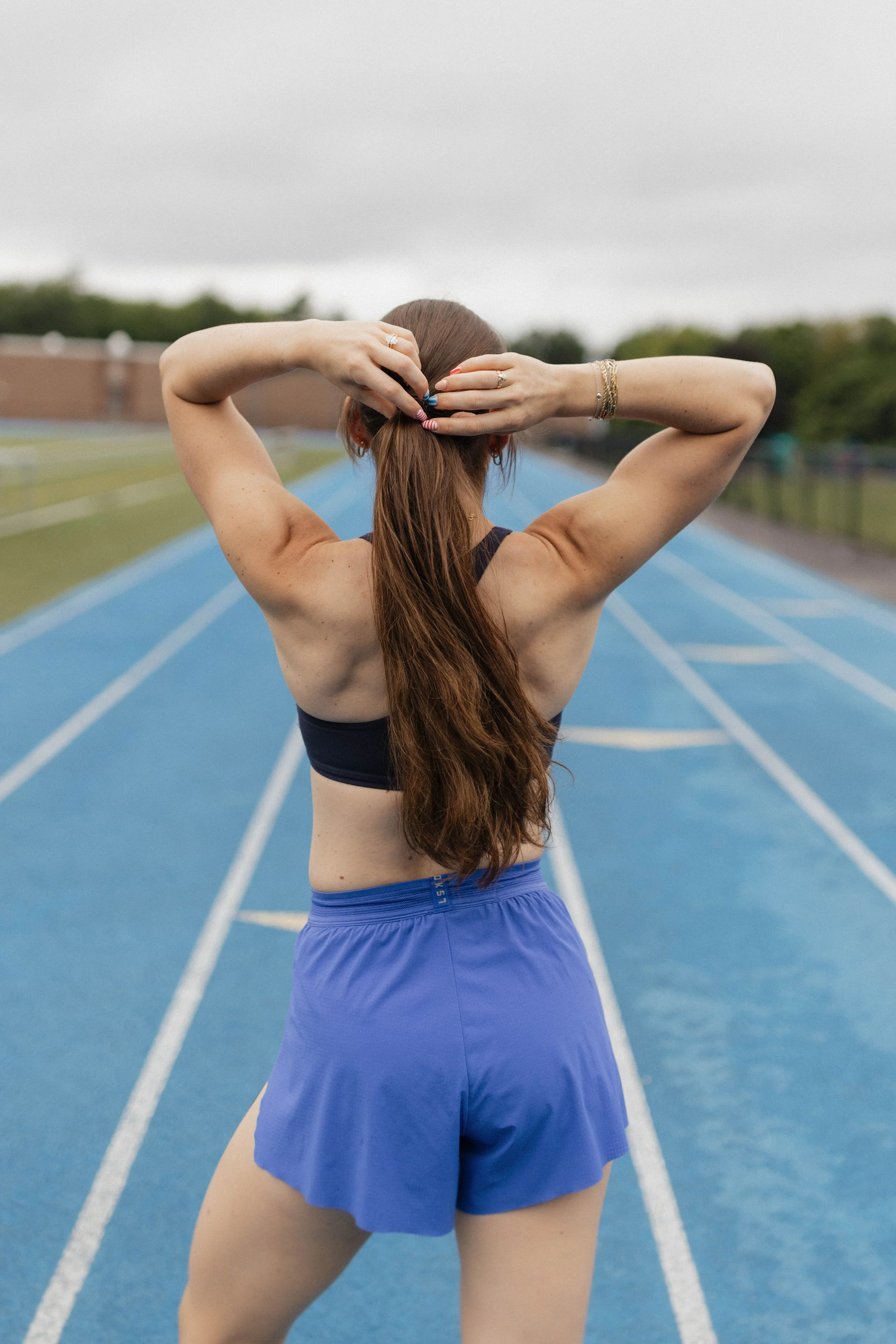 Back of a woman with long reddish hair wearing a black sports bra and blue shorts on a blue running track, stretching her arms behind her head. Overcast sky in the background.