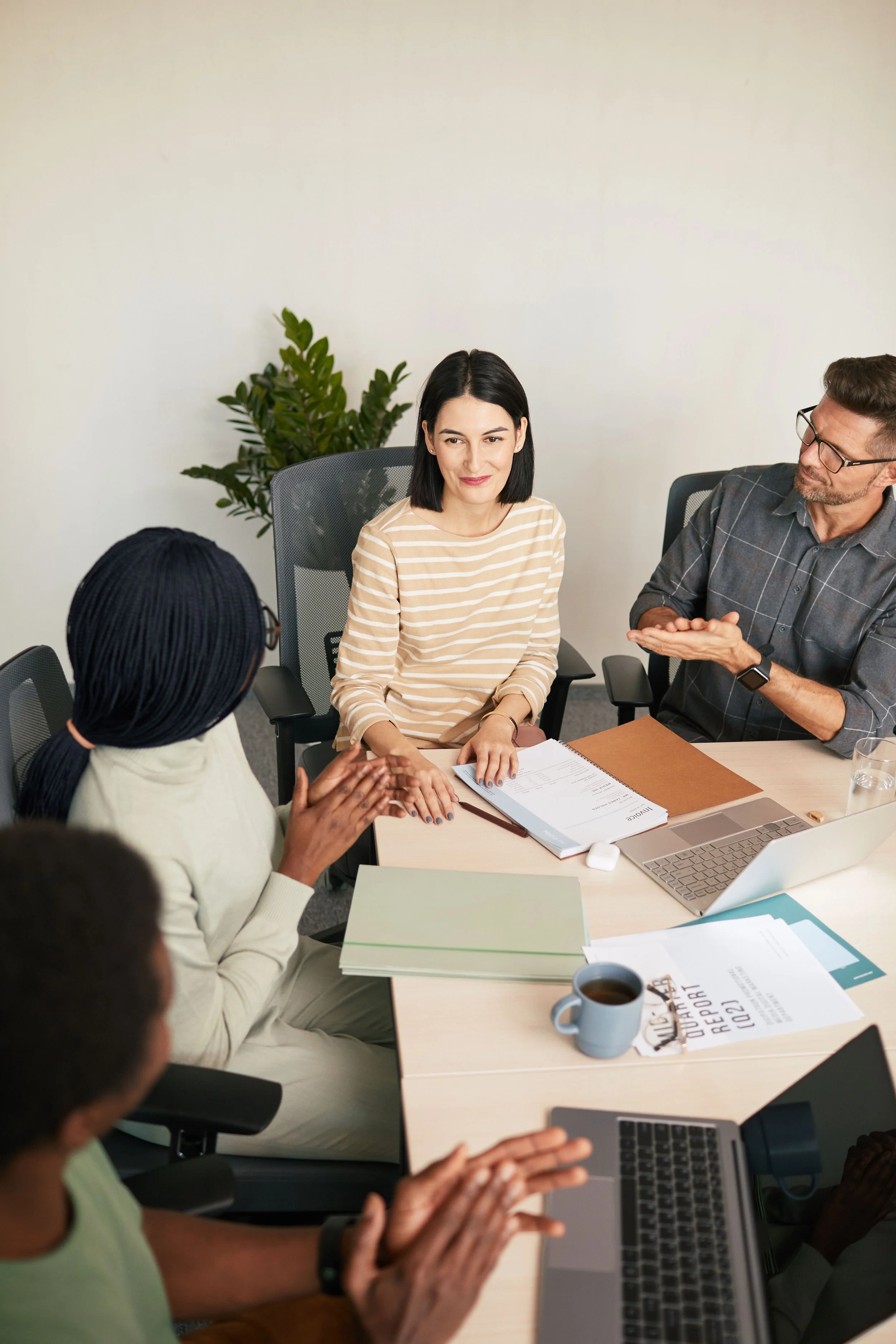 A diverse group of professionals sitting around a conference table engaged in a discussion, with documents, laptops, and a coffee mug on the table.
