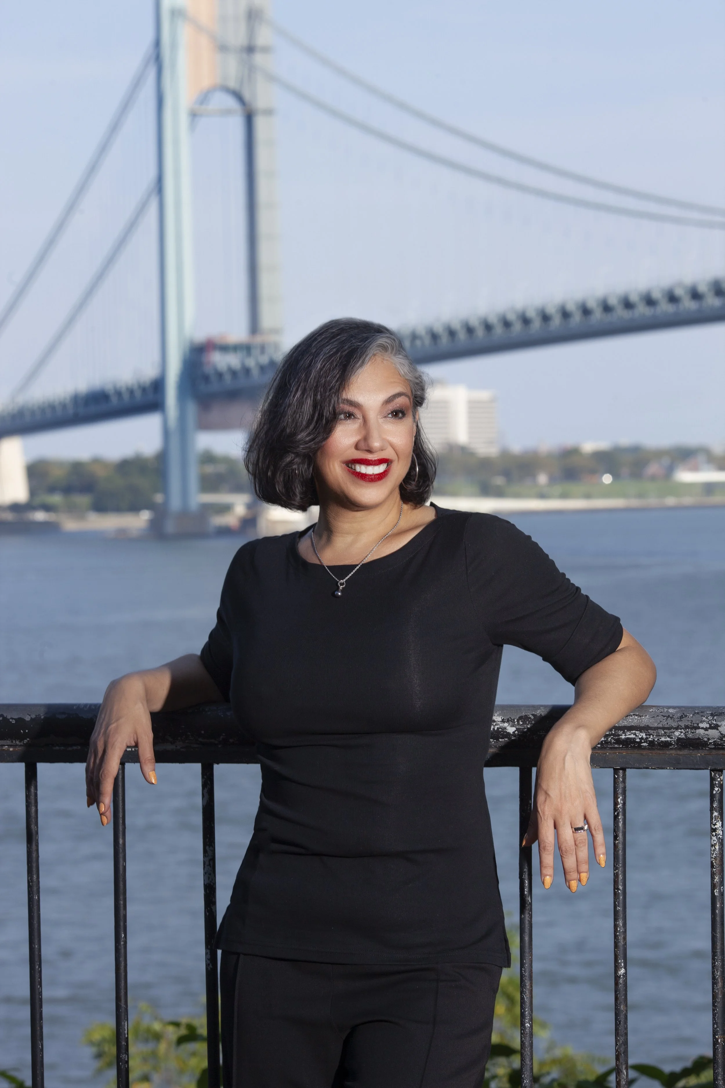 A woman with black and gray hair wearing a black dress and red lipstick standing by a black railing near a river, with a large bridge and city buildings in the background.