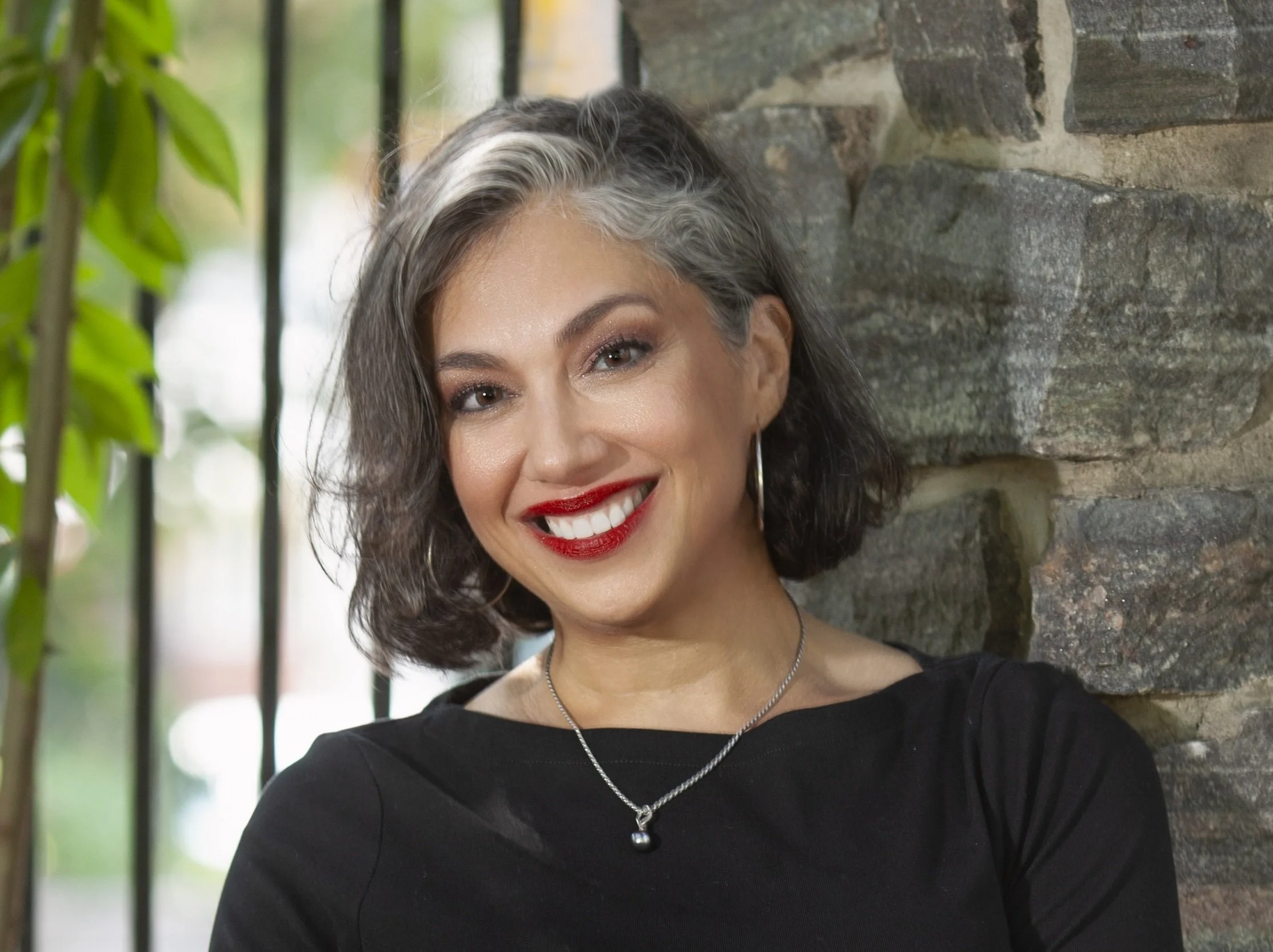 A smiling woman with shoulder-length curly hair, wearing a black top, a necklace with a pendant, and red lipstick, standing outdoors near a stone wall and green plants.
