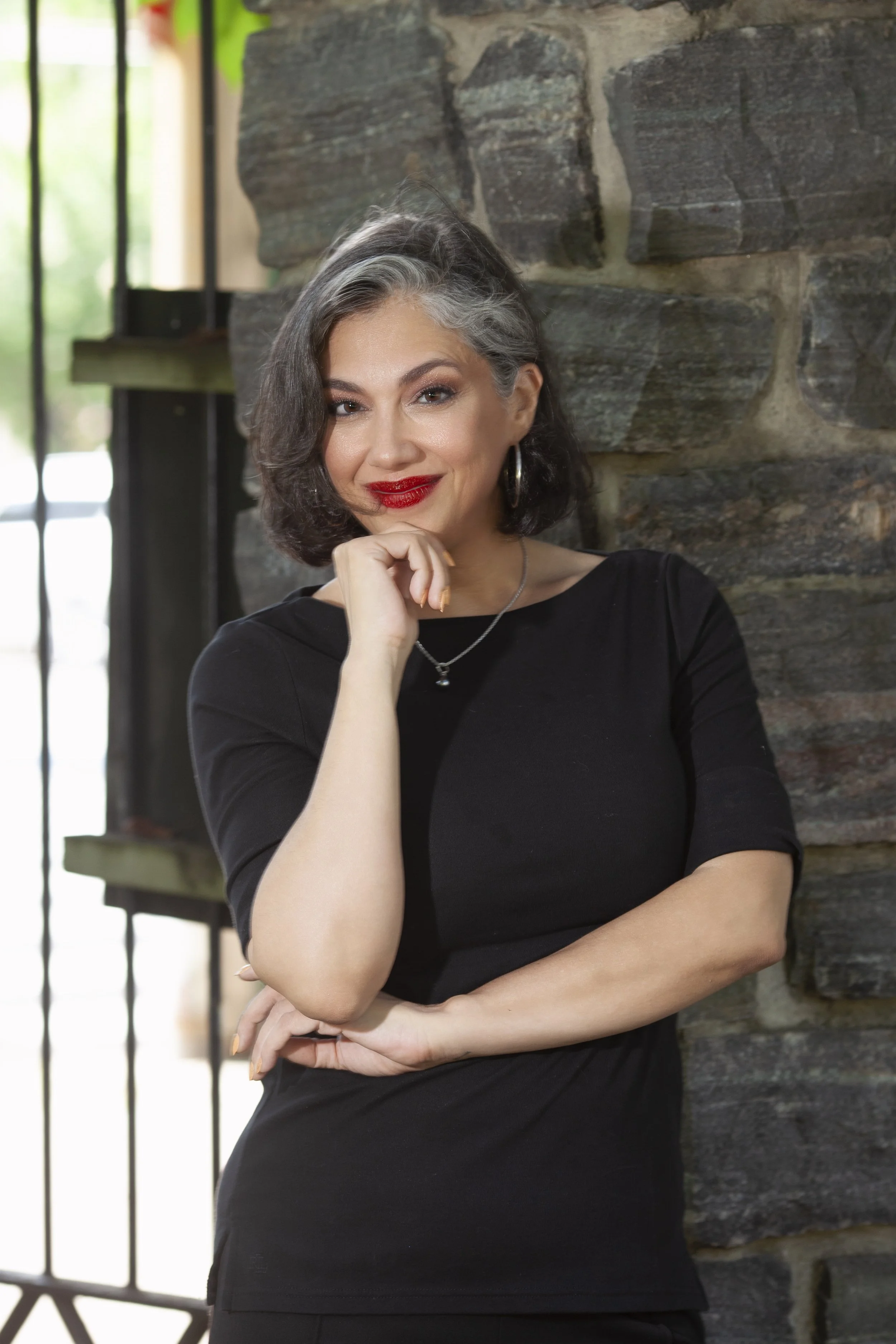 A woman with short, gray-streaked dark hair and red lipstick, wearing a black top, silver hoop earrings, and a necklace, standing with her hand resting on her chin in front of a stone wall and a window with metal bars.