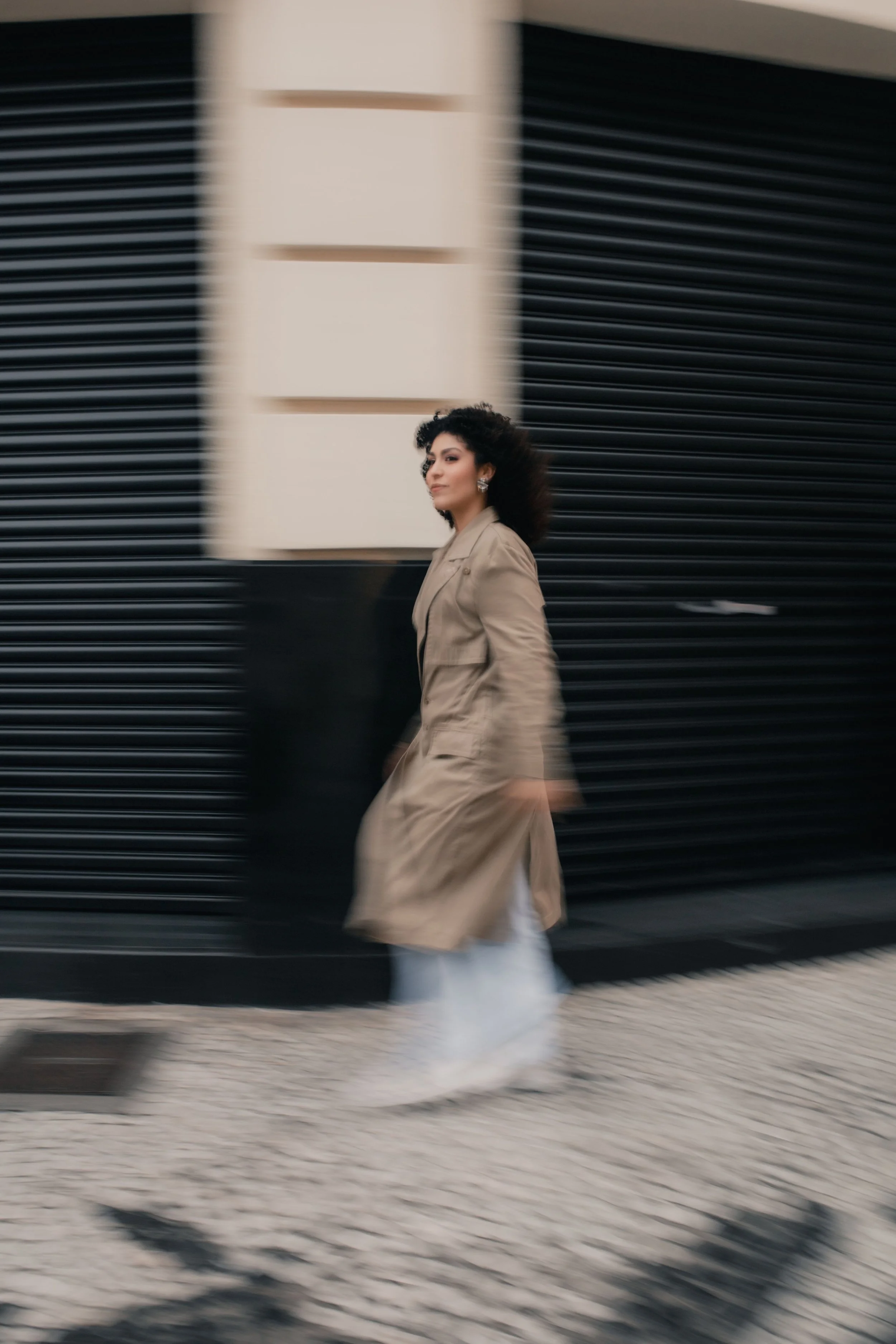 Woman walking past black slatted wall, wearing beige trench coat and white pants, with blurred motion.