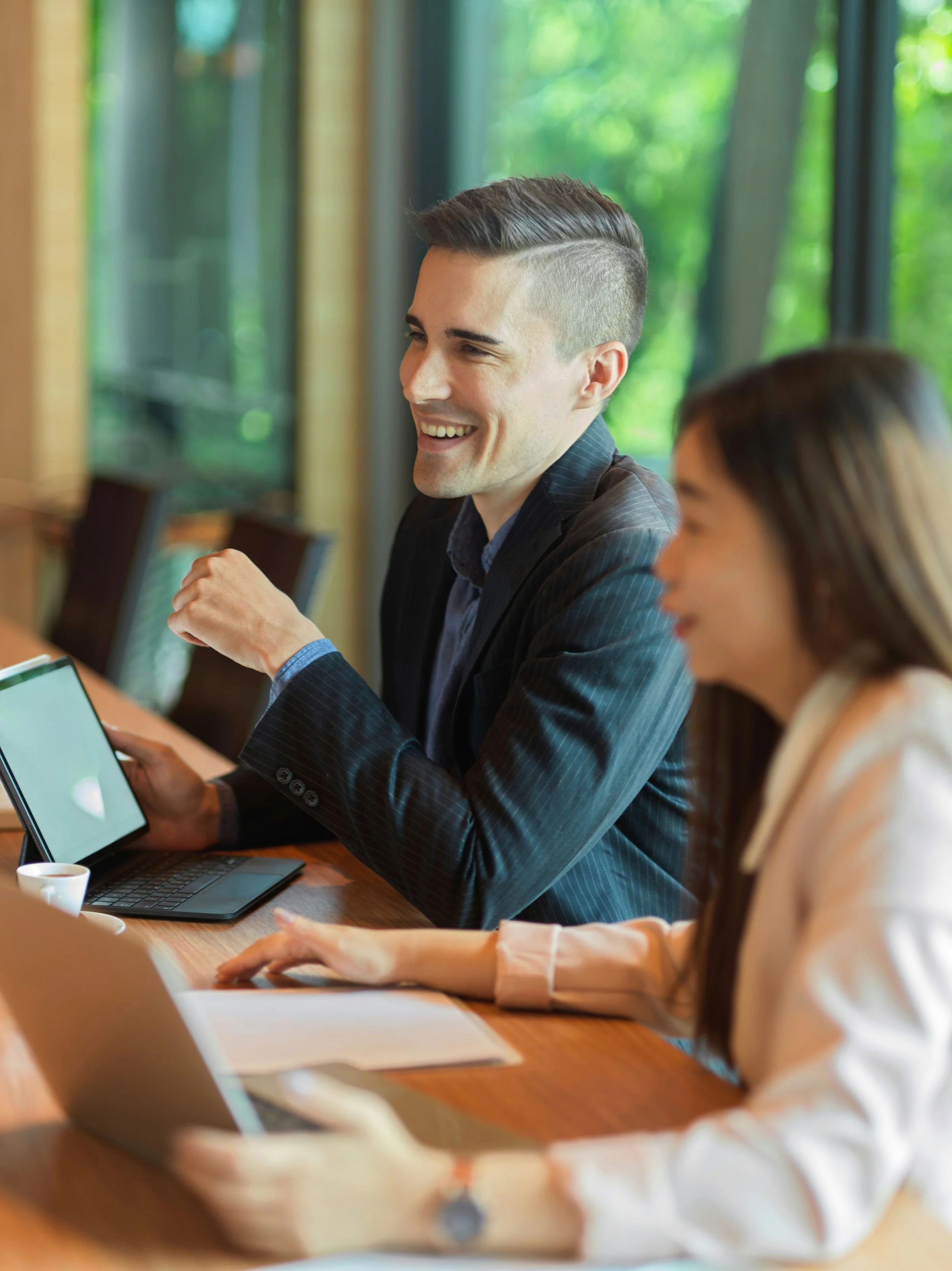 A young man and a woman with long brown hair sitting at a wooden table in a meeting room with large windows showing green trees. They are smiling, and the man is holding a tablet while the woman has a laptop in front of her.