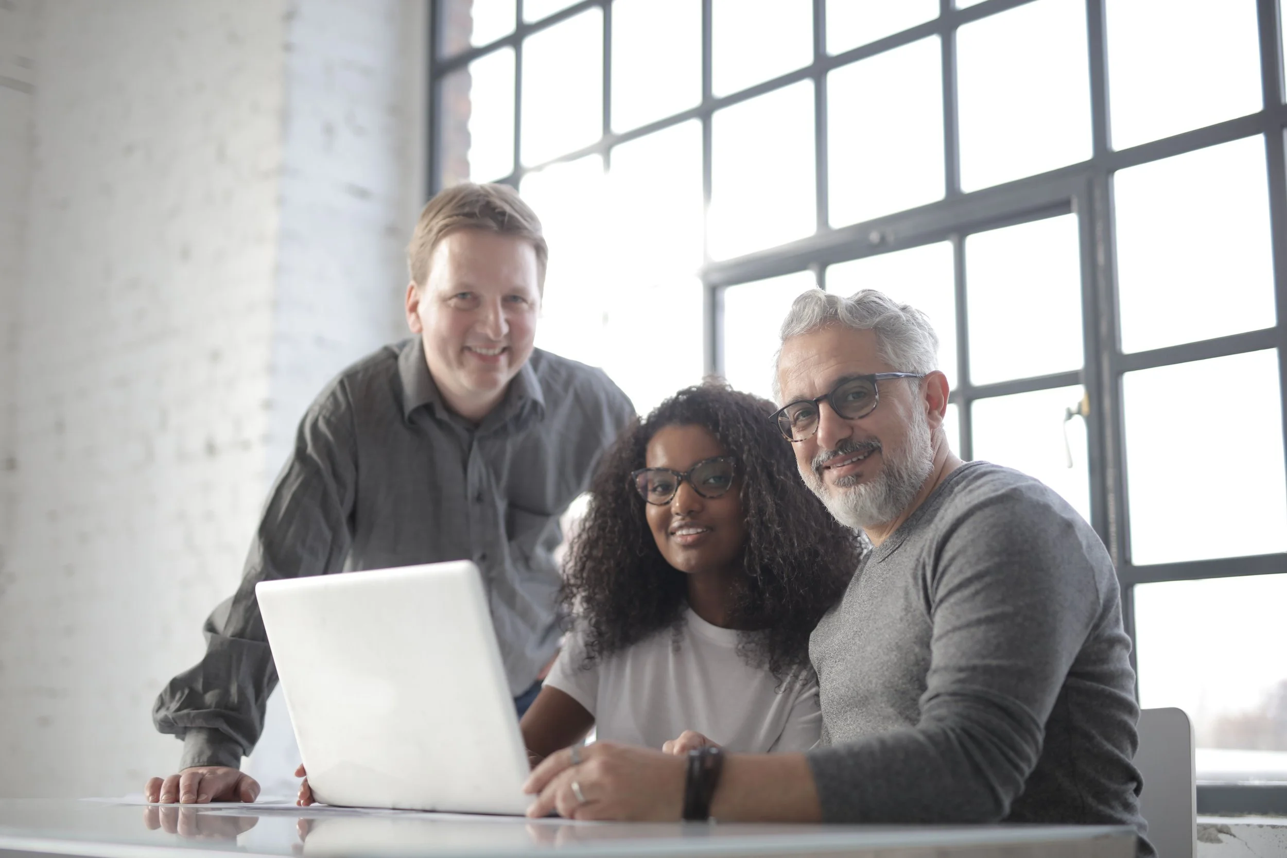 Three diverse people gathered around a laptop in a modern office with large windows, smiling and looking at the camera.