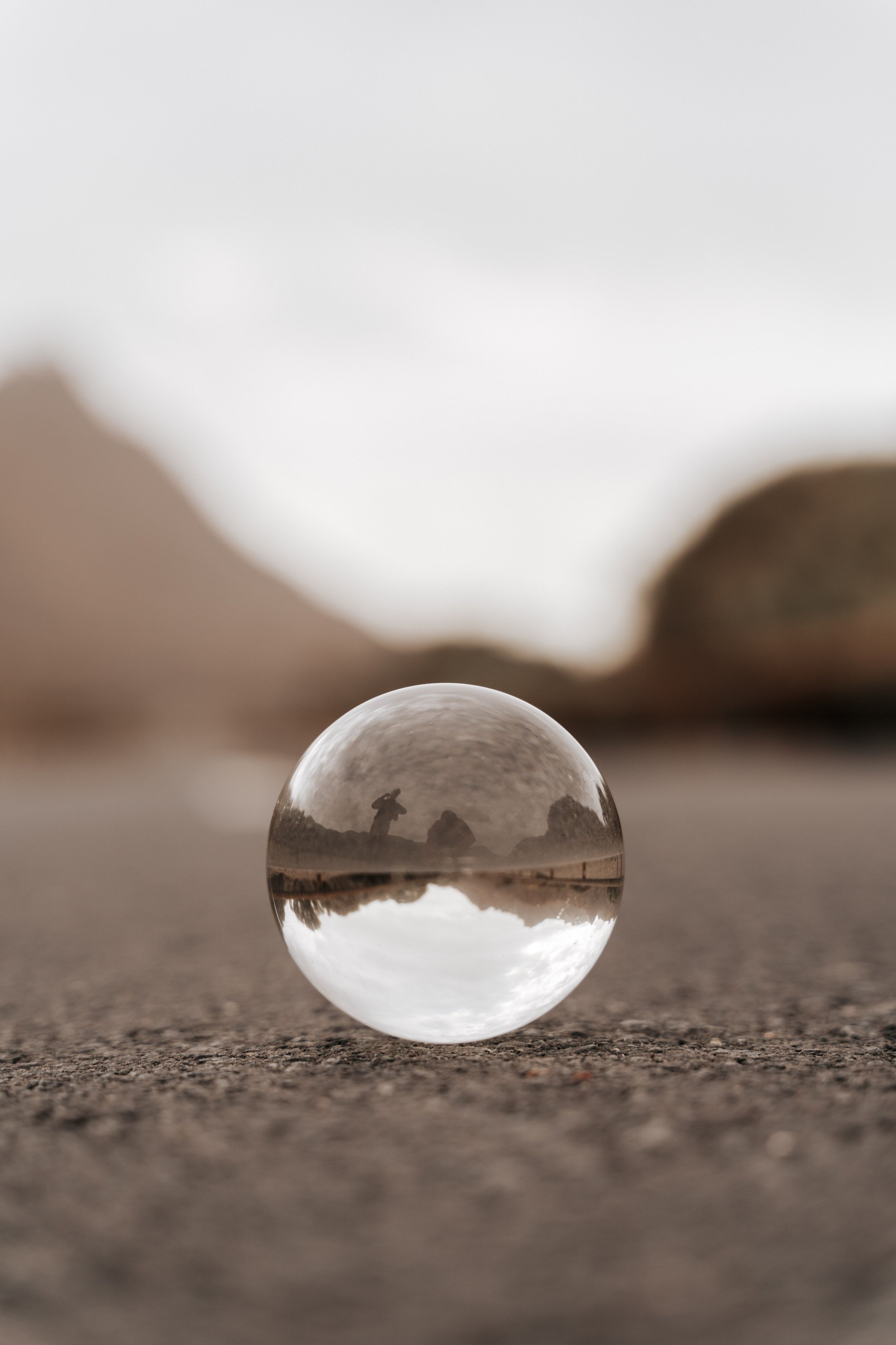 A transparent glass sphere sits on a textured gray surface, reflecting a cloudy sky and silhouettes of two people, with blurred hills in the background.