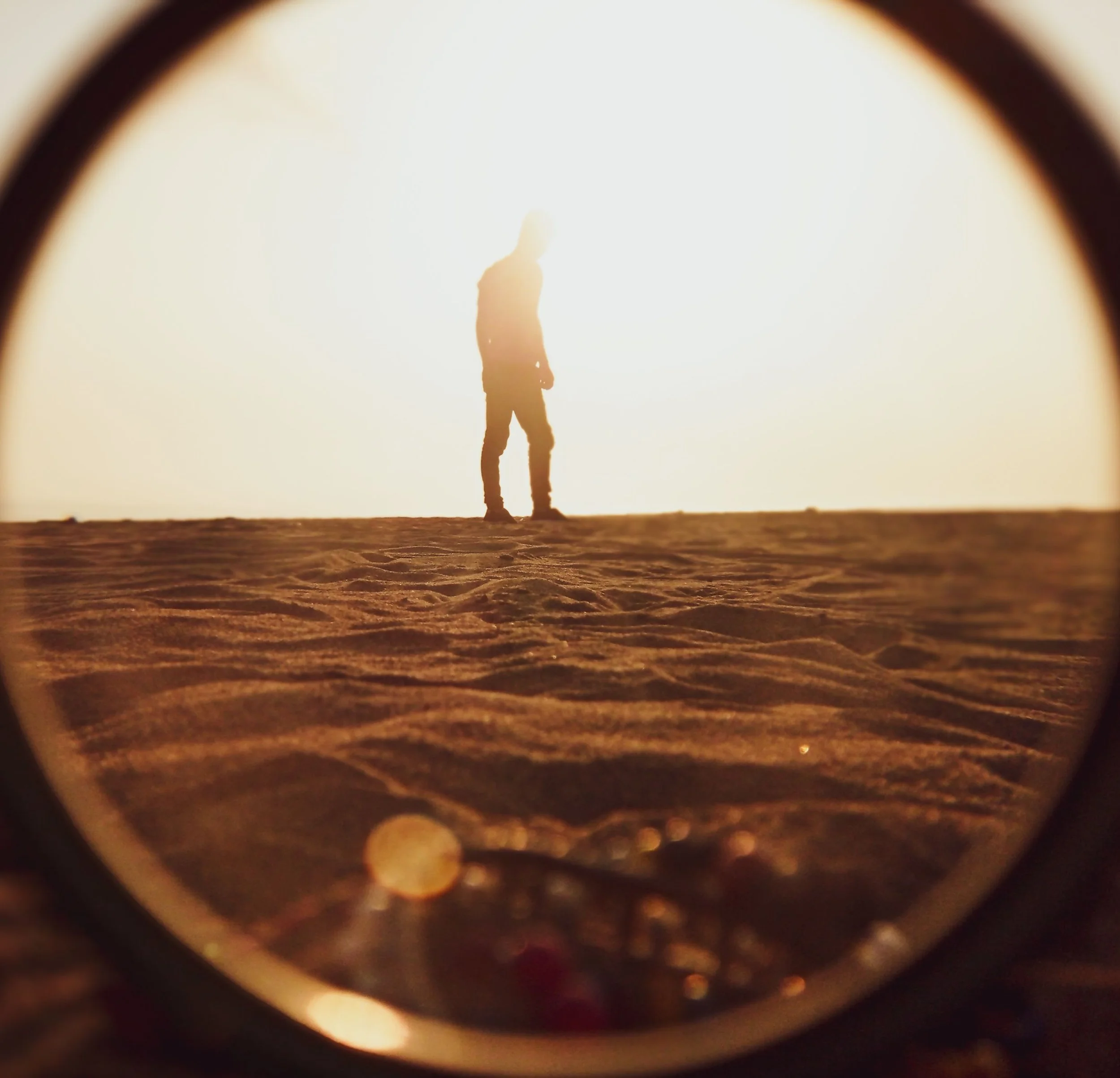 A person standing on a beach during sunset, viewed through a tunnel or pipe with a circular opening.