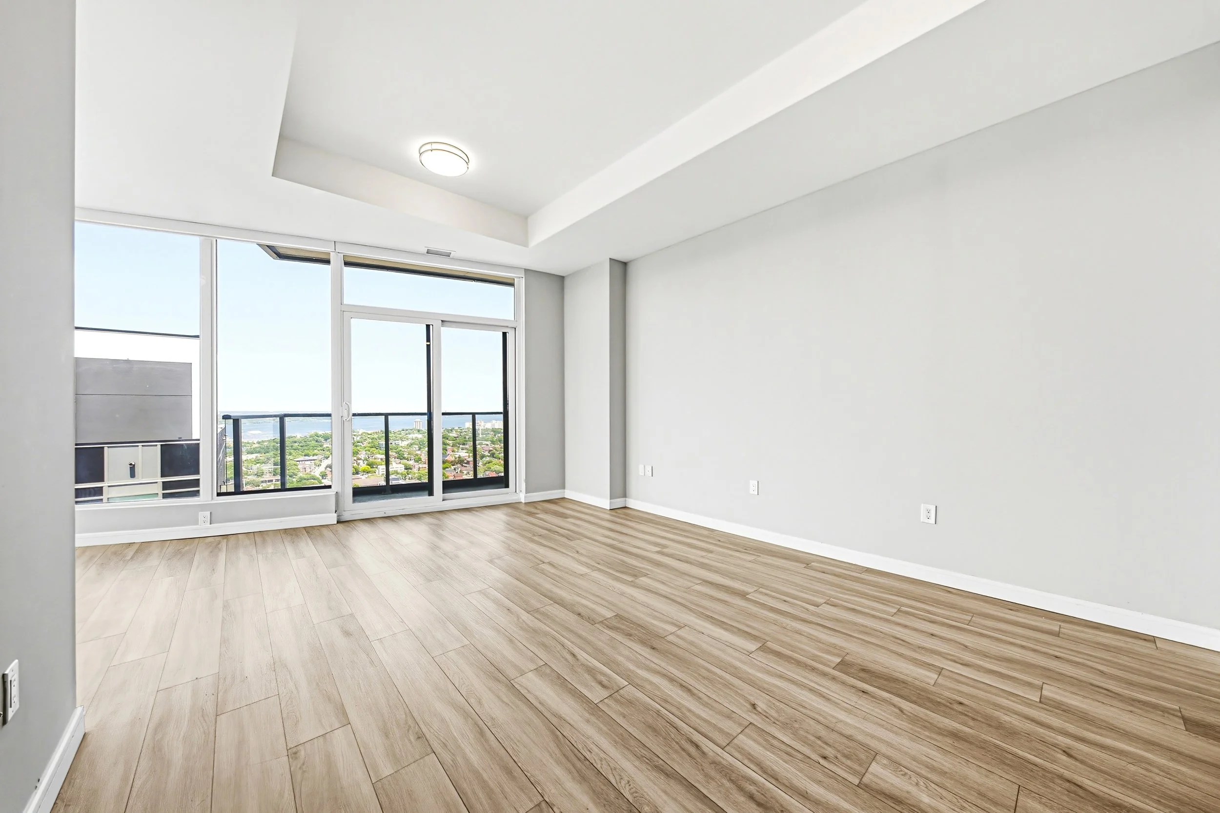 Empty room with large windows overlooking a cityscape, white walls, and light wood flooring.