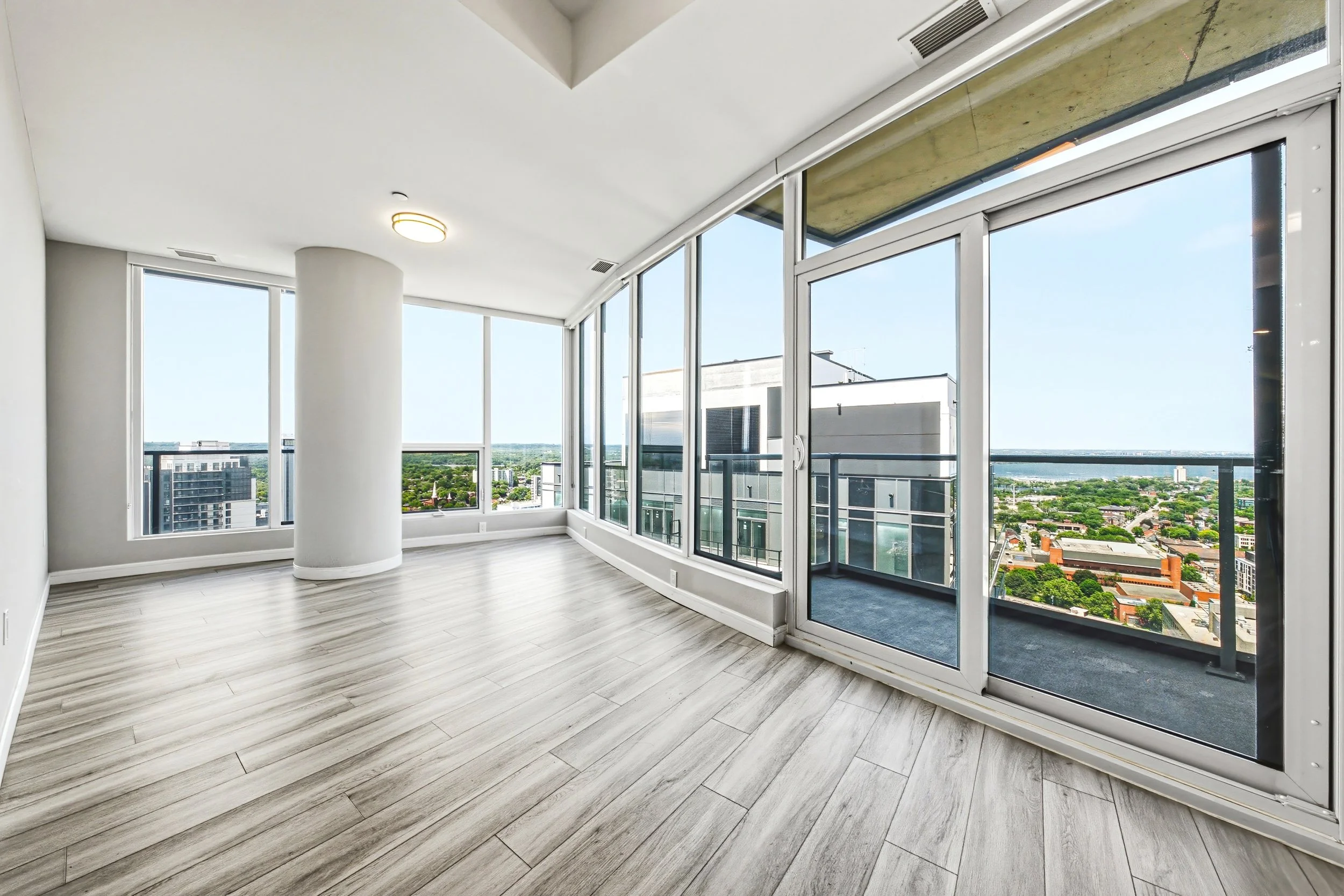 Modern high-rise apartment with large windows offering city views, light-colored wood flooring, and a minimalist ceiling light.