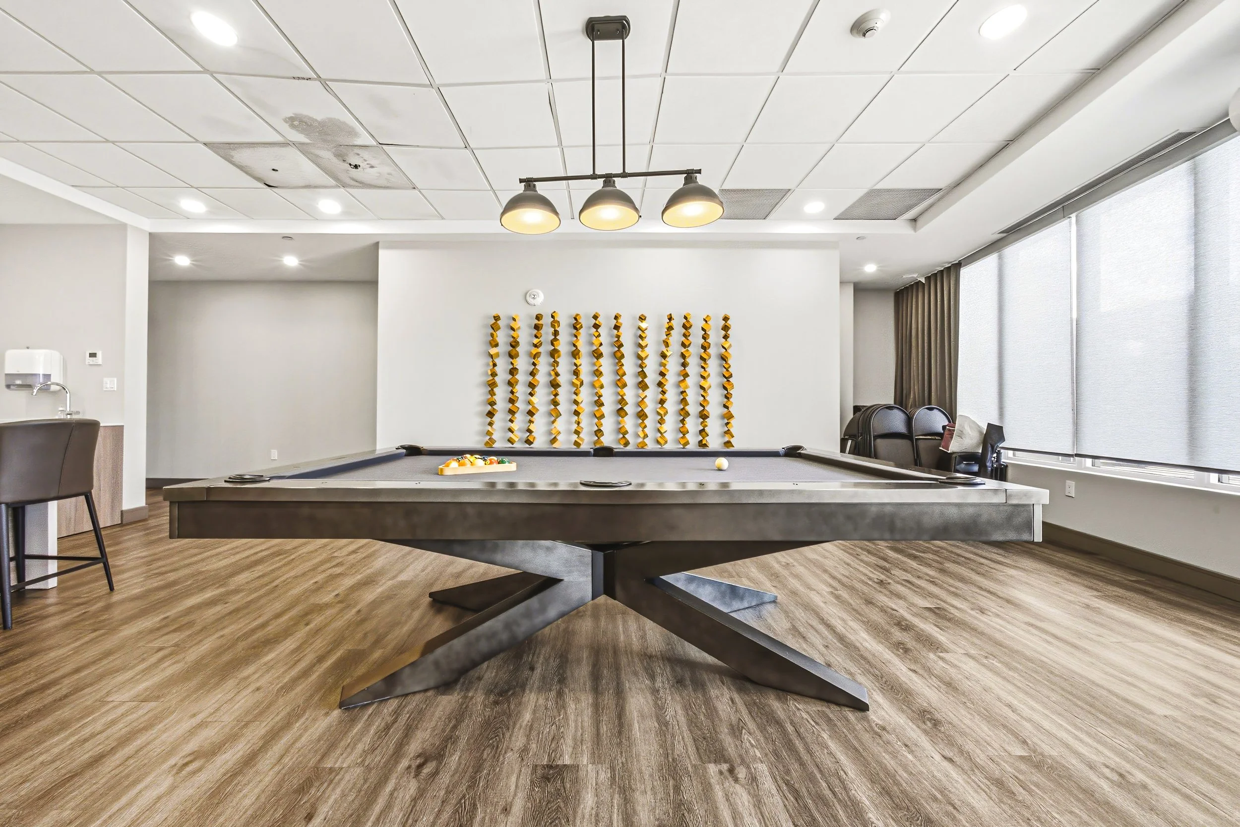 Indoor room with a pool table, wall-mounted yellow decorative objects, chairs, large windows with blinds, and ceiling lights.