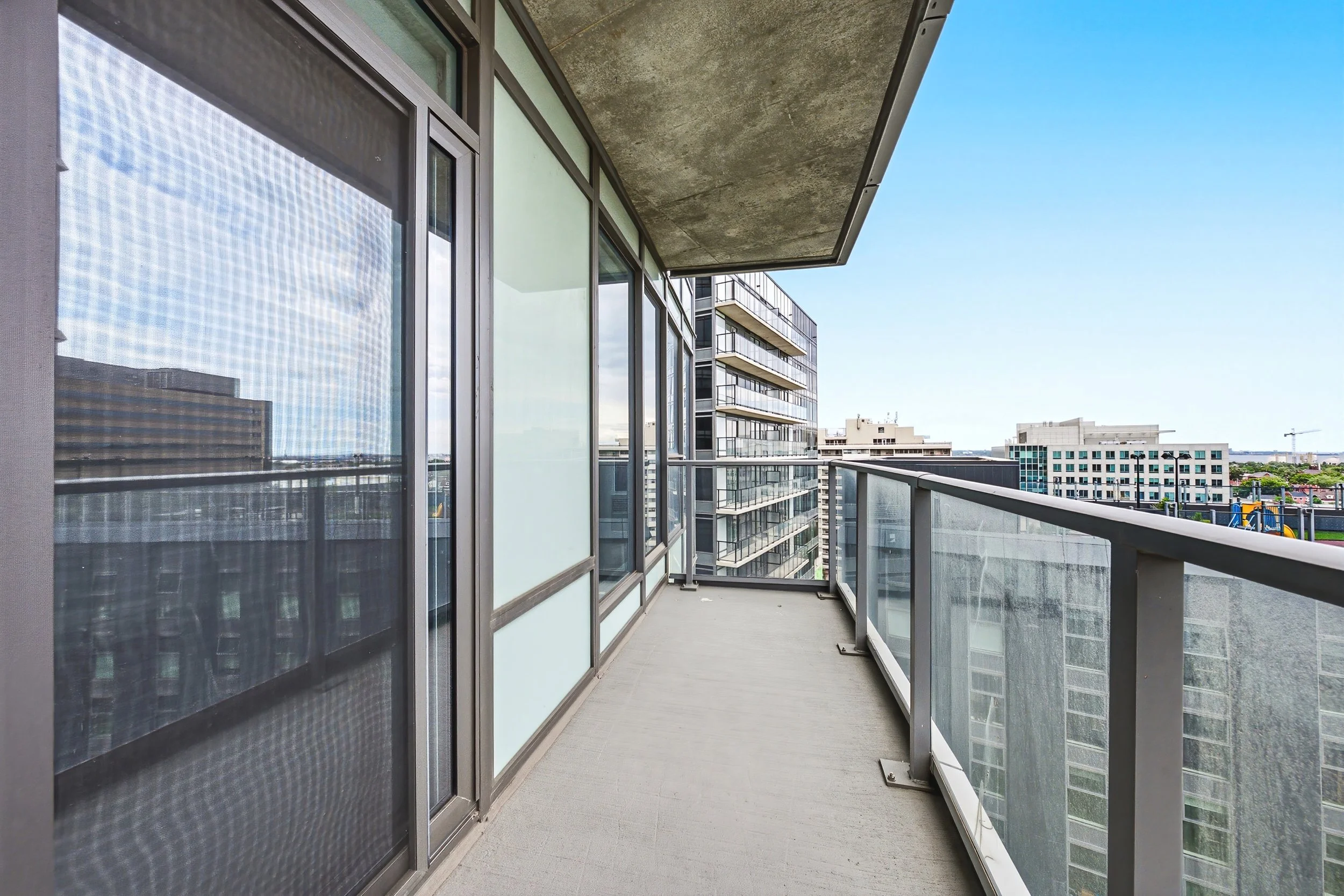 An empty high-rise apartment balcony with glass and metal railing, overlooking other buildings and a playground in the distance under a blue sky.